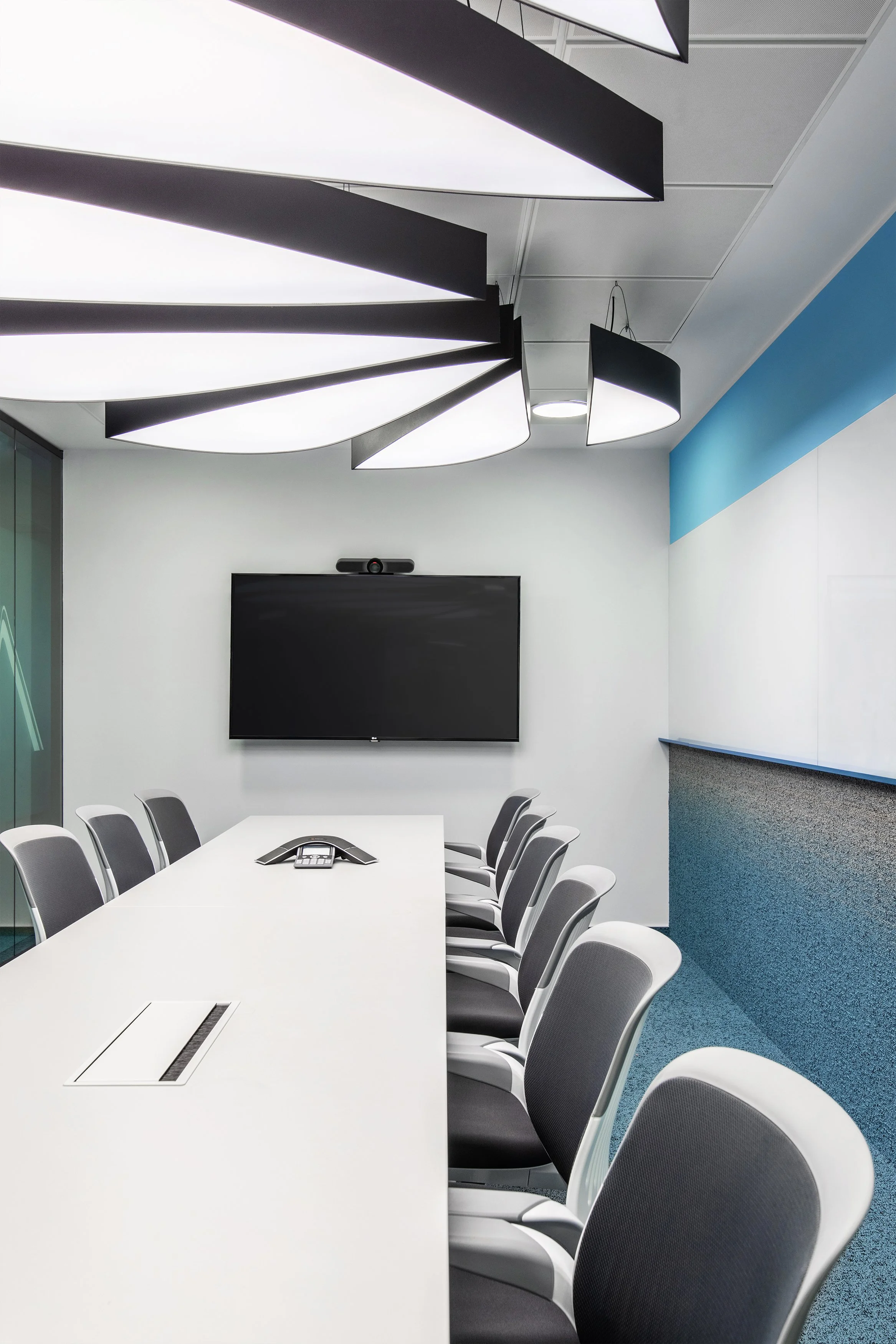 Empty modern conference room with a long white table, gray chairs, a black wall-mounted television, a video camera above the TV, and contemporary lighting fixtures on the ceiling.