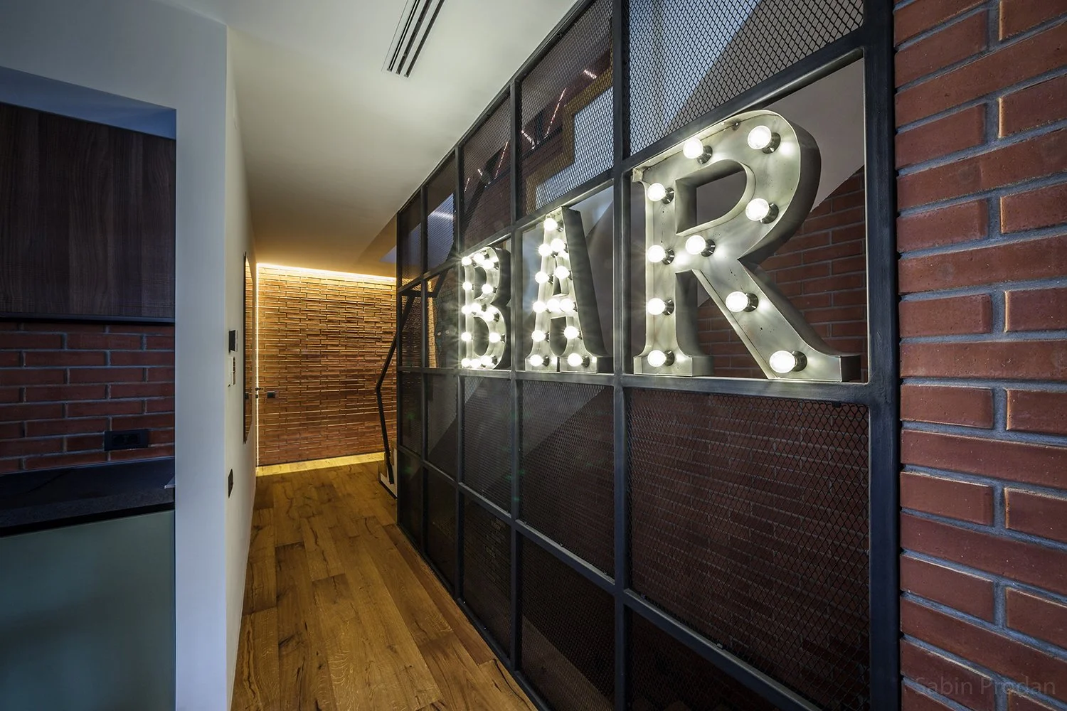 Interior hallway featuring a lit sign spelling 'BAR' with letter bulbs mounted on a metal grid wall, brick wall to the side, hardwood floor, and modern decor.