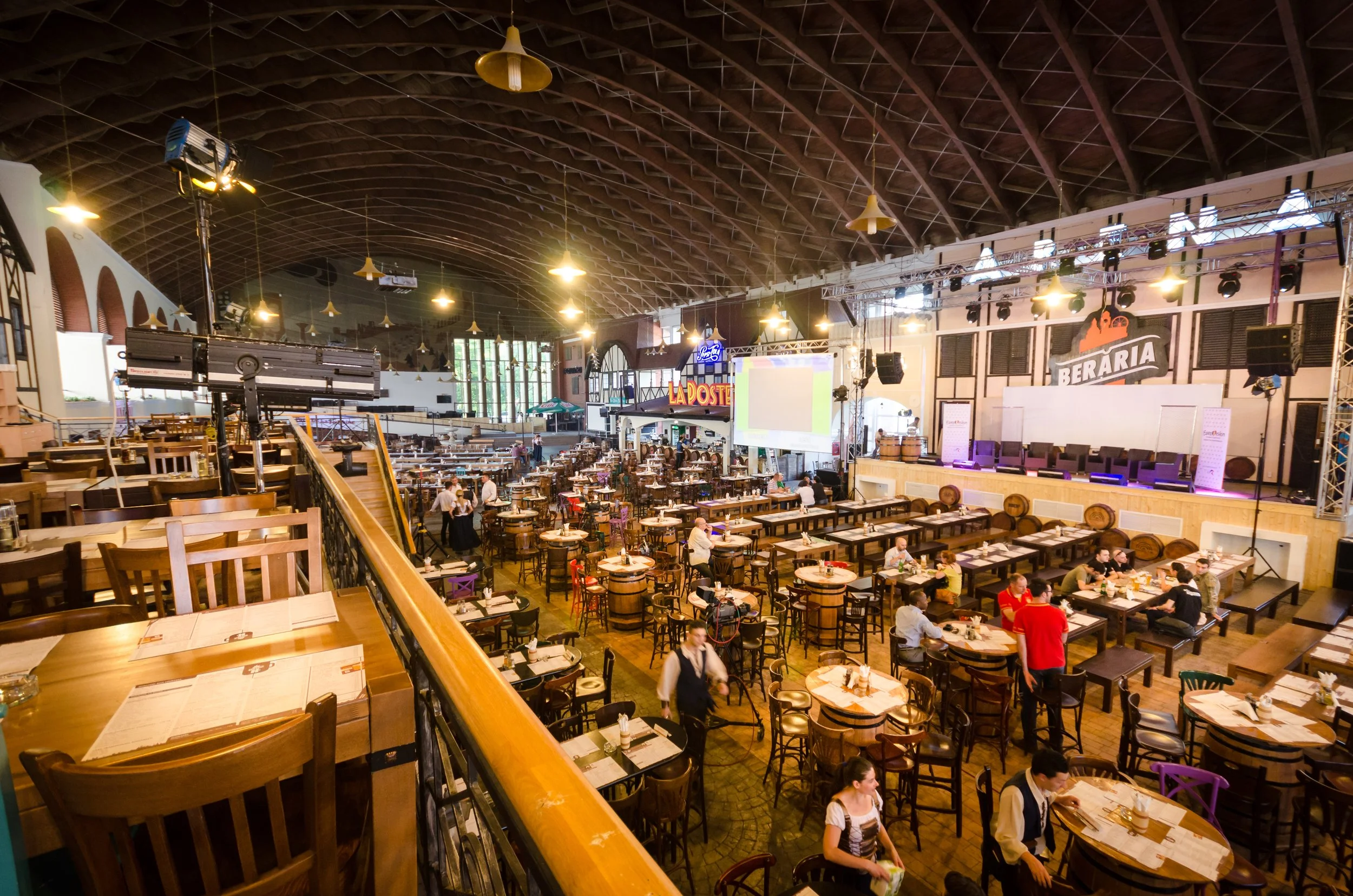 Wide view of a bustling restaurant or event space with wooden tables and chairs, a stage, and large screens, illuminated with warm lighting under a high, arched ceiling.