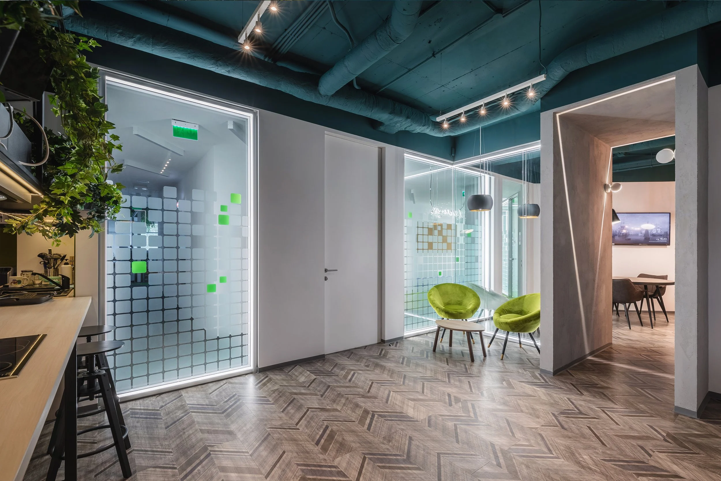 Modern office lounge area with two green velvet chairs, small wooden table, decorative lighting, and glass walls with green and white square decals, hardwood floor, and a partial view of a kitchen area.