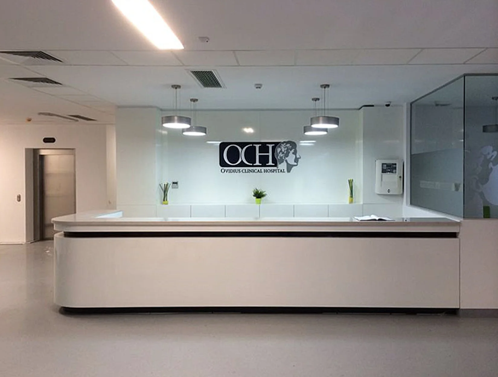 Hospital reception area with white curved counter, logo of Ovidius Clinical Hospital on the wall, three overhead lights, potted plants, a white panel with medical supplies, and an elevator in the background.
