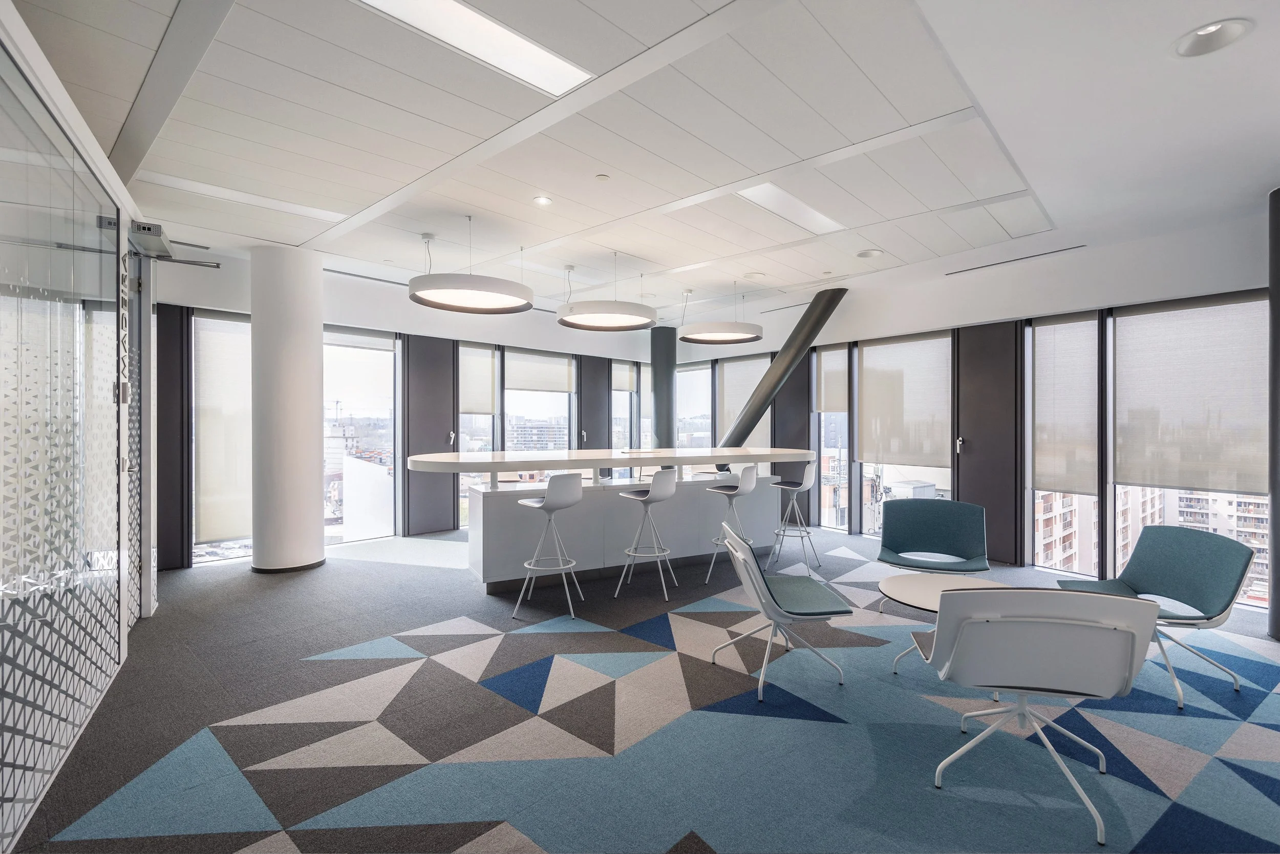 Modern office lounge with large windows, round ceiling lights, a high table with bar stools, and seating area with chairs and a small round table, featuring a geometric patterned carpet.