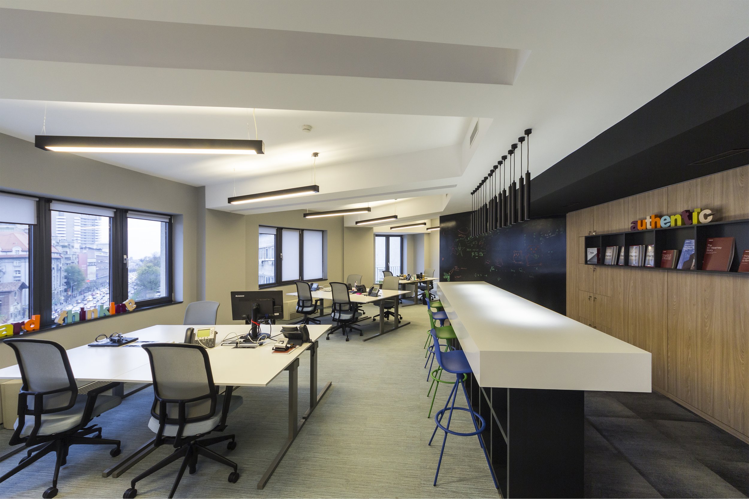 Modern office with multiple desks, chairs, computers, and a bar-height counter with colorful stools. Large windows with city views, black ceiling lights, and decorative bookshelves with colorful letters spelling 'authentic' on the wall.
