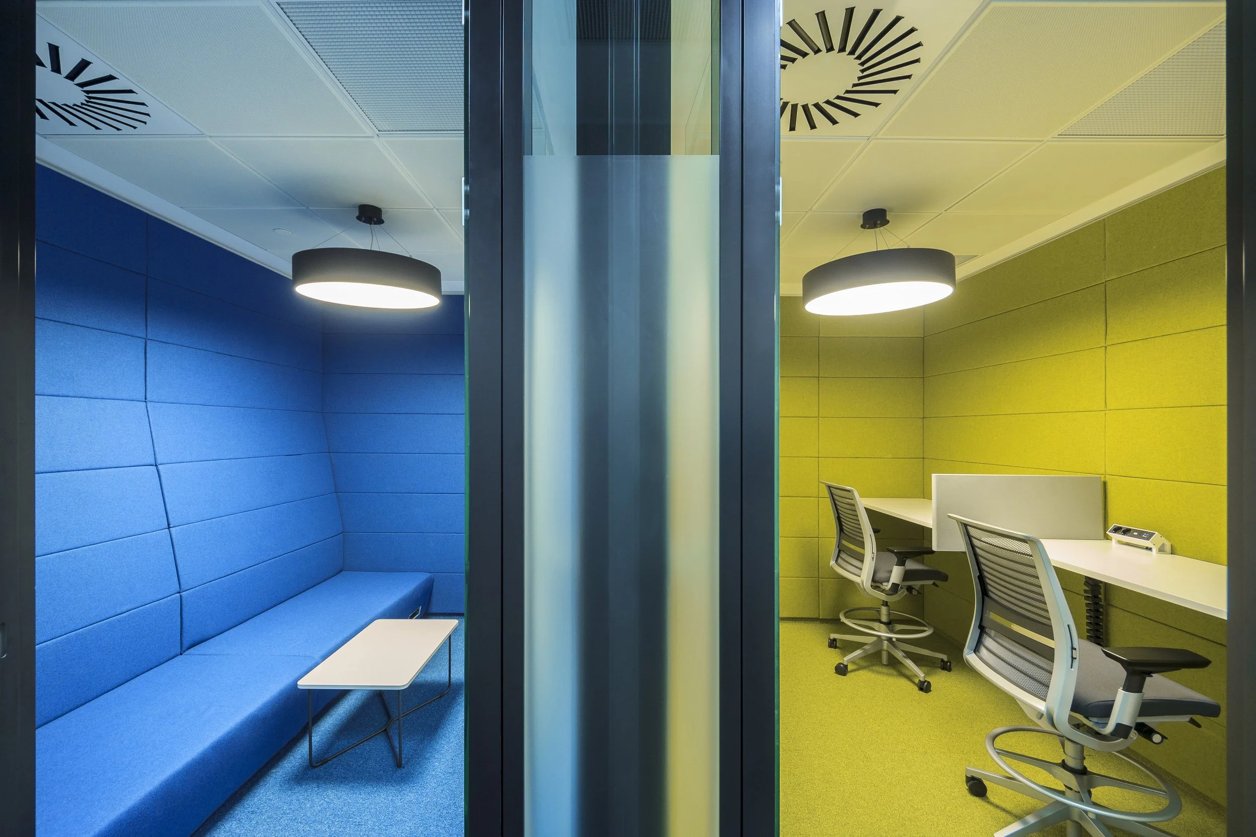 Two adjacent modern office booths, one with blue walls and seating, the other with yellow walls and desks, separated by glass and metal barriers.