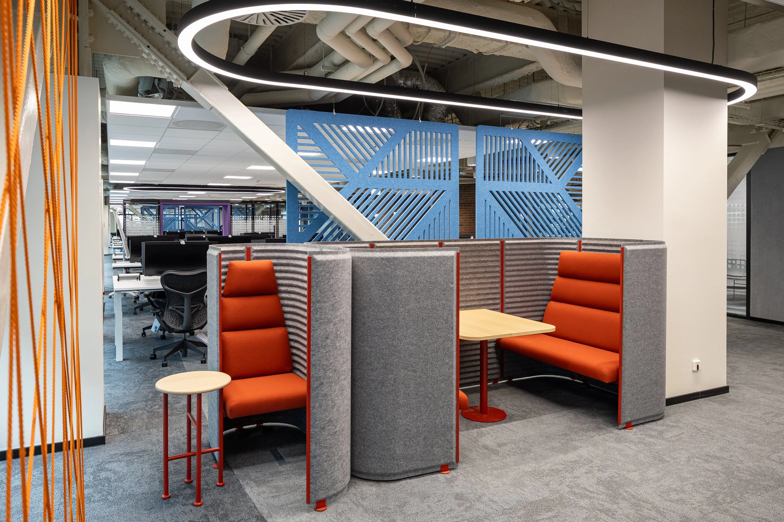 Cozy seating area with two orange cushioned booths separated by gray partitions and small wooden tables, in a modern office space with abstract blue wall panels and industrial ceiling design.