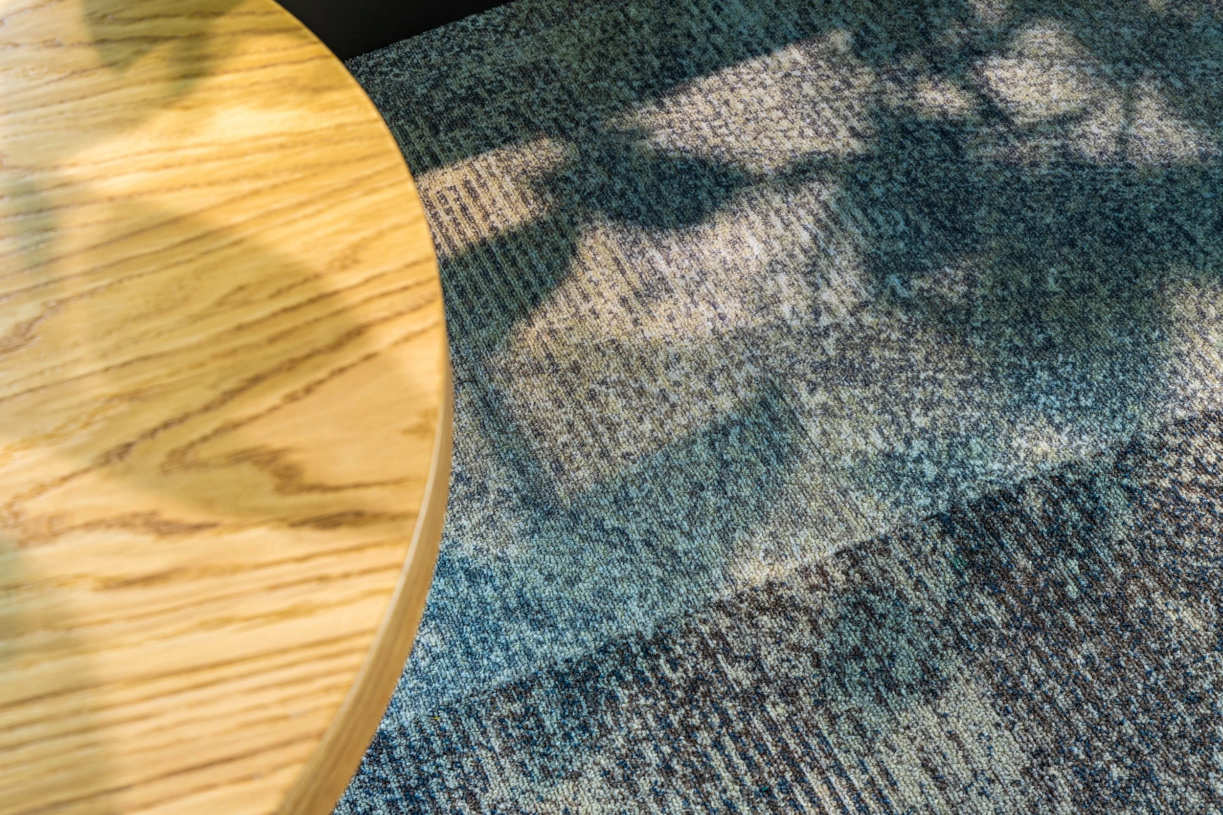 Close-up of a wooden table with visible grain, positioned next to a textured carpet surface with shadows cast across it.