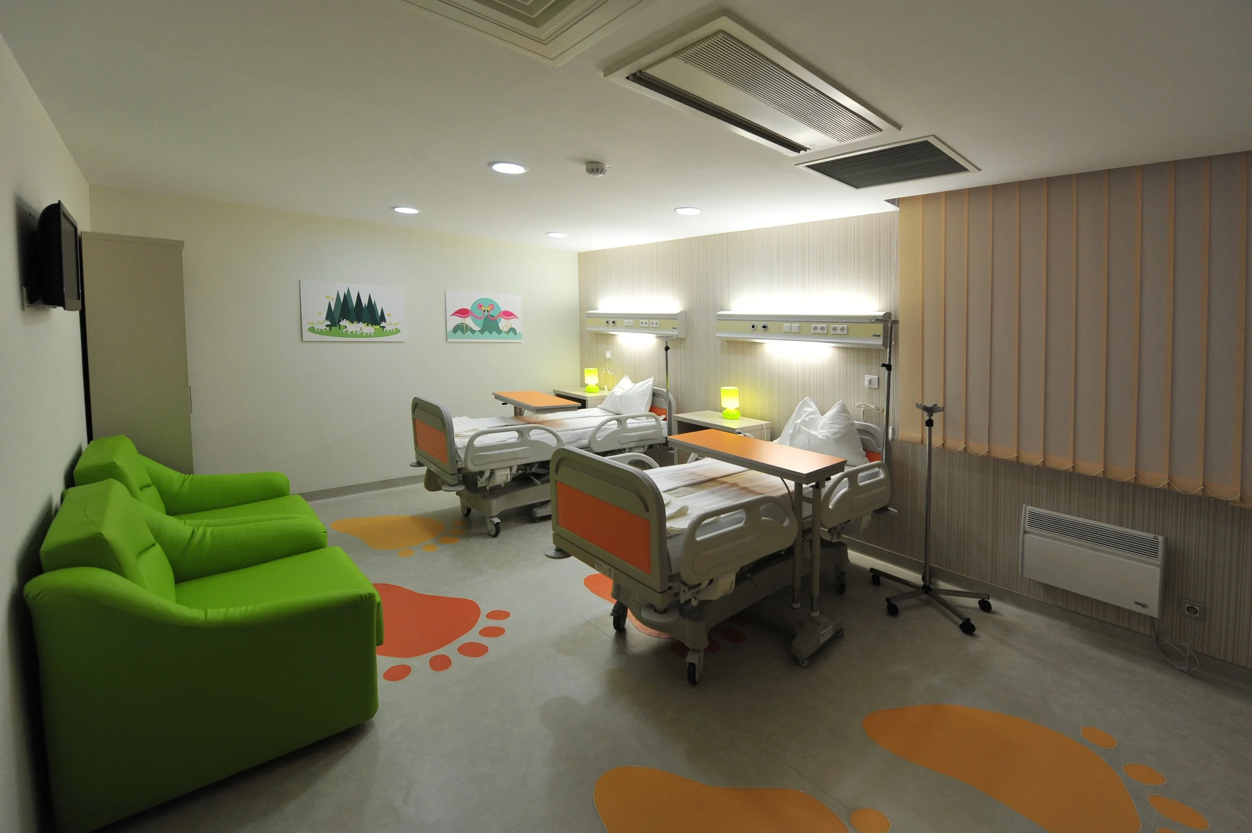 A clean, brightly lit hospital room with two beds, each with an orange desk, green lamps, and medical equipment. Two green chairs are on the left side, and pastel-colored artwork is on the wall.