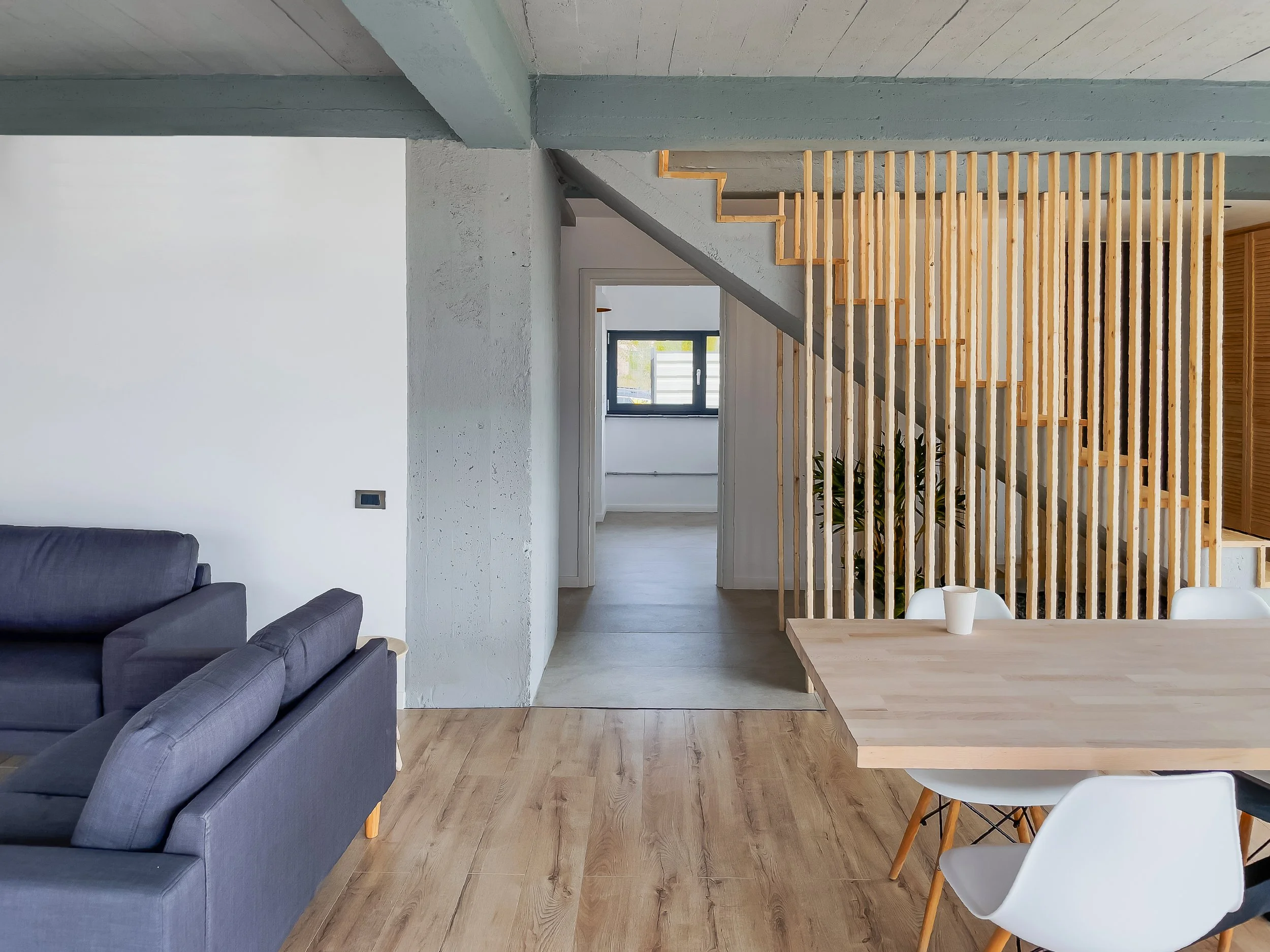 Modern living room with gray sofas, wooden flooring, and a partial view of a dining table with white chairs, separated by wooden slats, and a doorway leading to a room with a window.