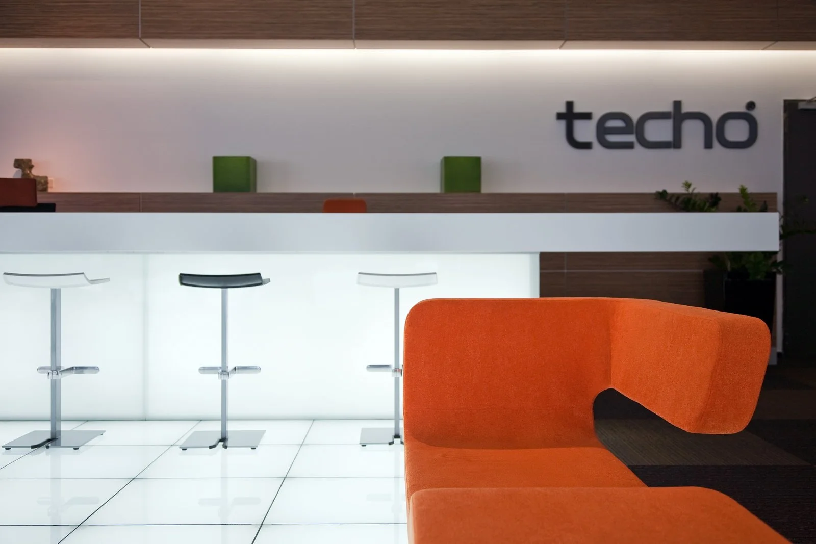 Modern office reception area with an orange curved sofa, a white illuminated front desk, and three tall bar stools with black, white, and black seats in front of it. The background has a white wall with the logo "techo" and a wooden panel with two green decorative boxes and plants.