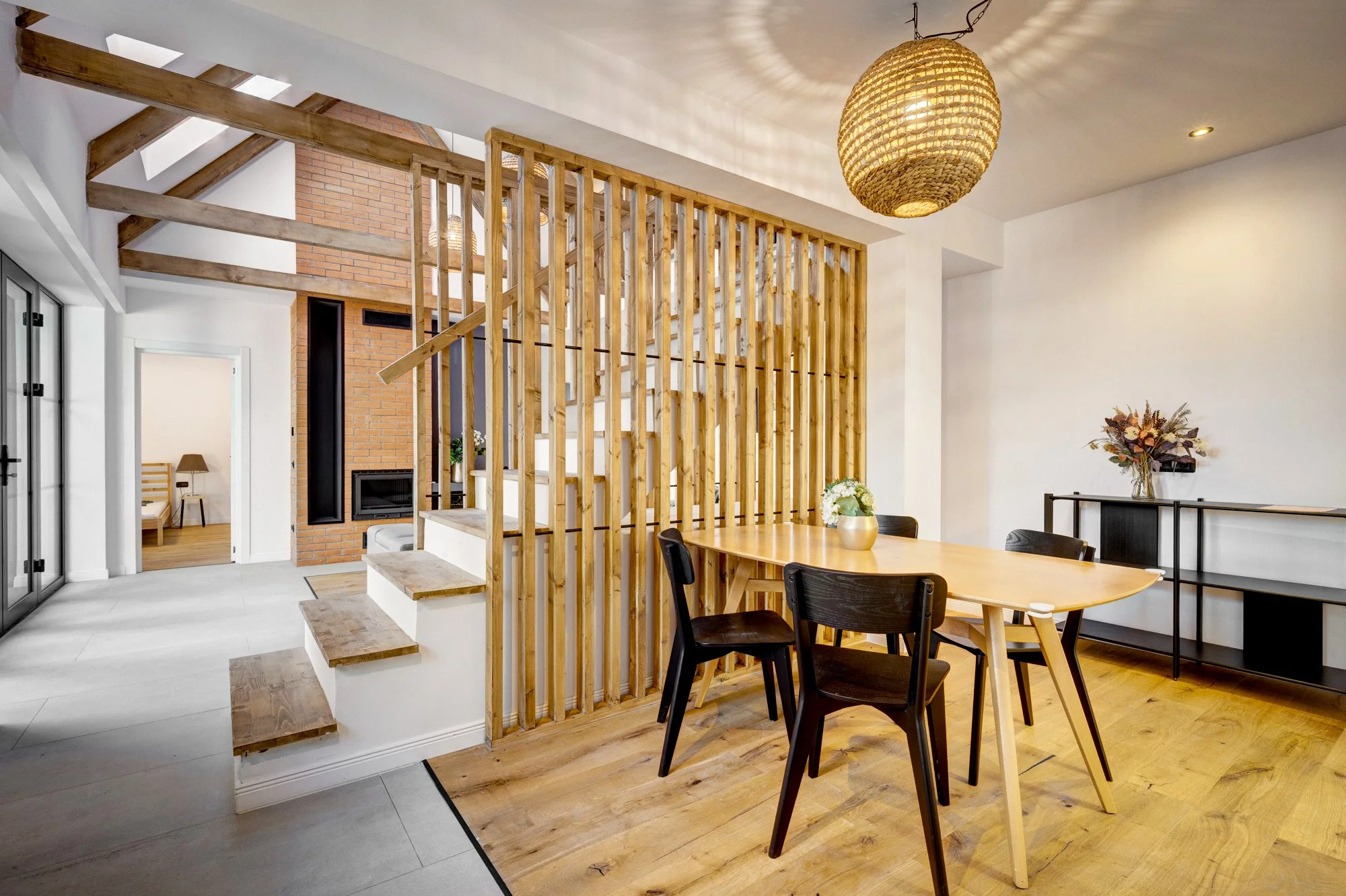 Open dining area with wooden table and black chairs, minimalist decor, wood flooring, wooden slat room divider, chandelier, white walls, doors leading to other rooms, and a fireplace area with brick wall.