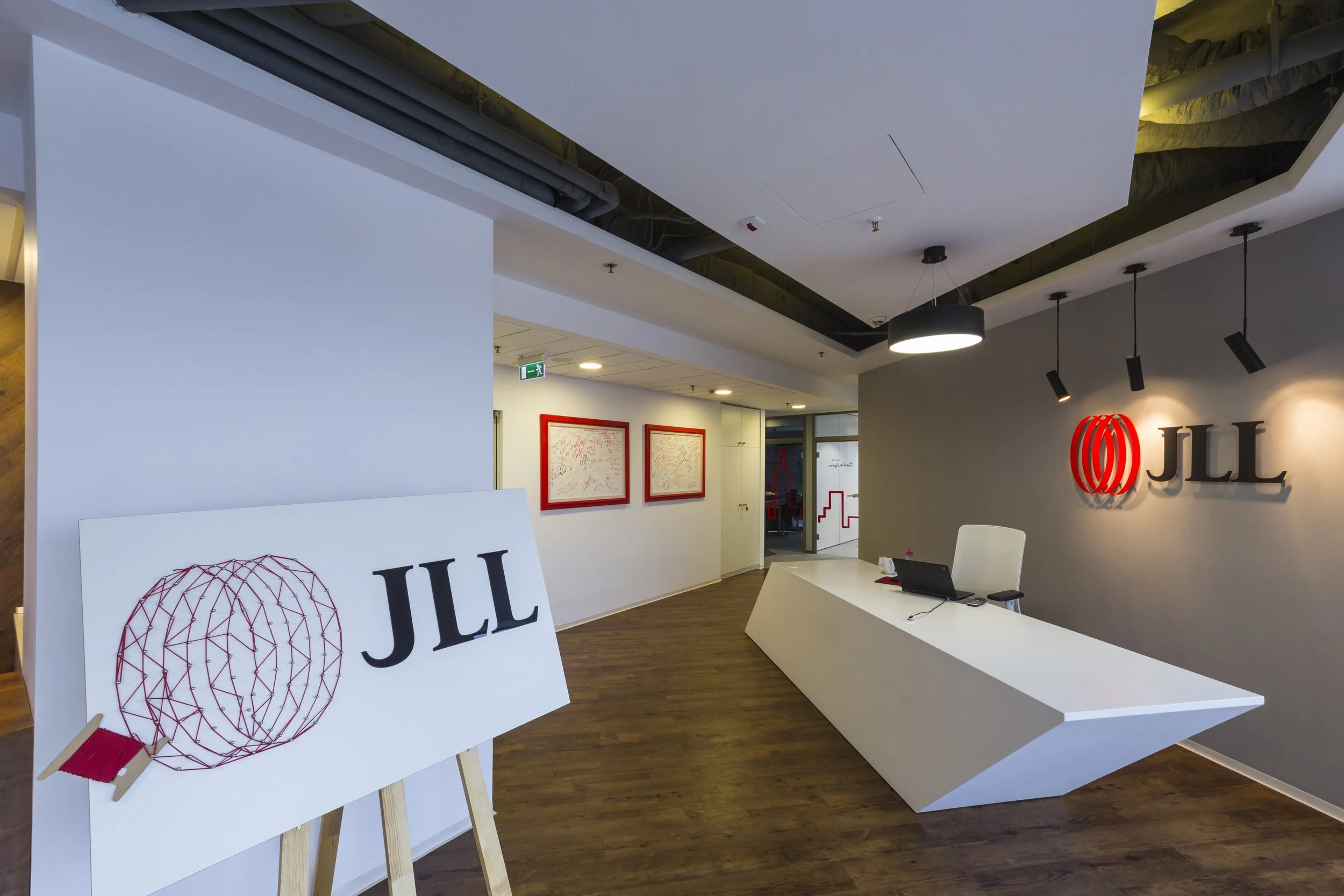 Reception area of JLL office with a white desk, a laptop, and JLL logo on the wall, illuminated by ceiling lights, with framed artwork and a doorway in the background.