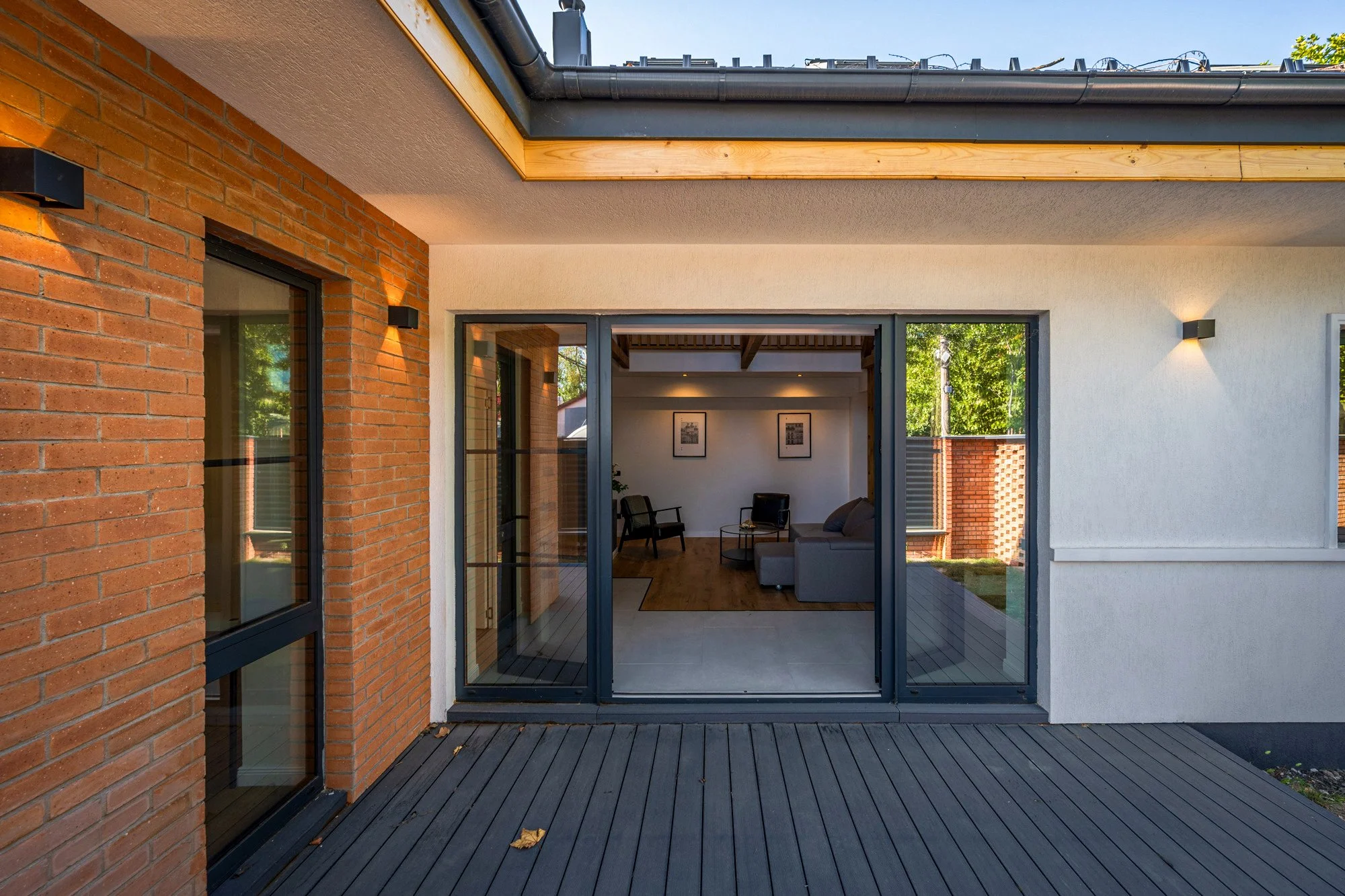 View of a modern house exterior with a sliding glass door opening into a living room, with a brick wall on the left and a white wall on the right, and a wooden deck outside.