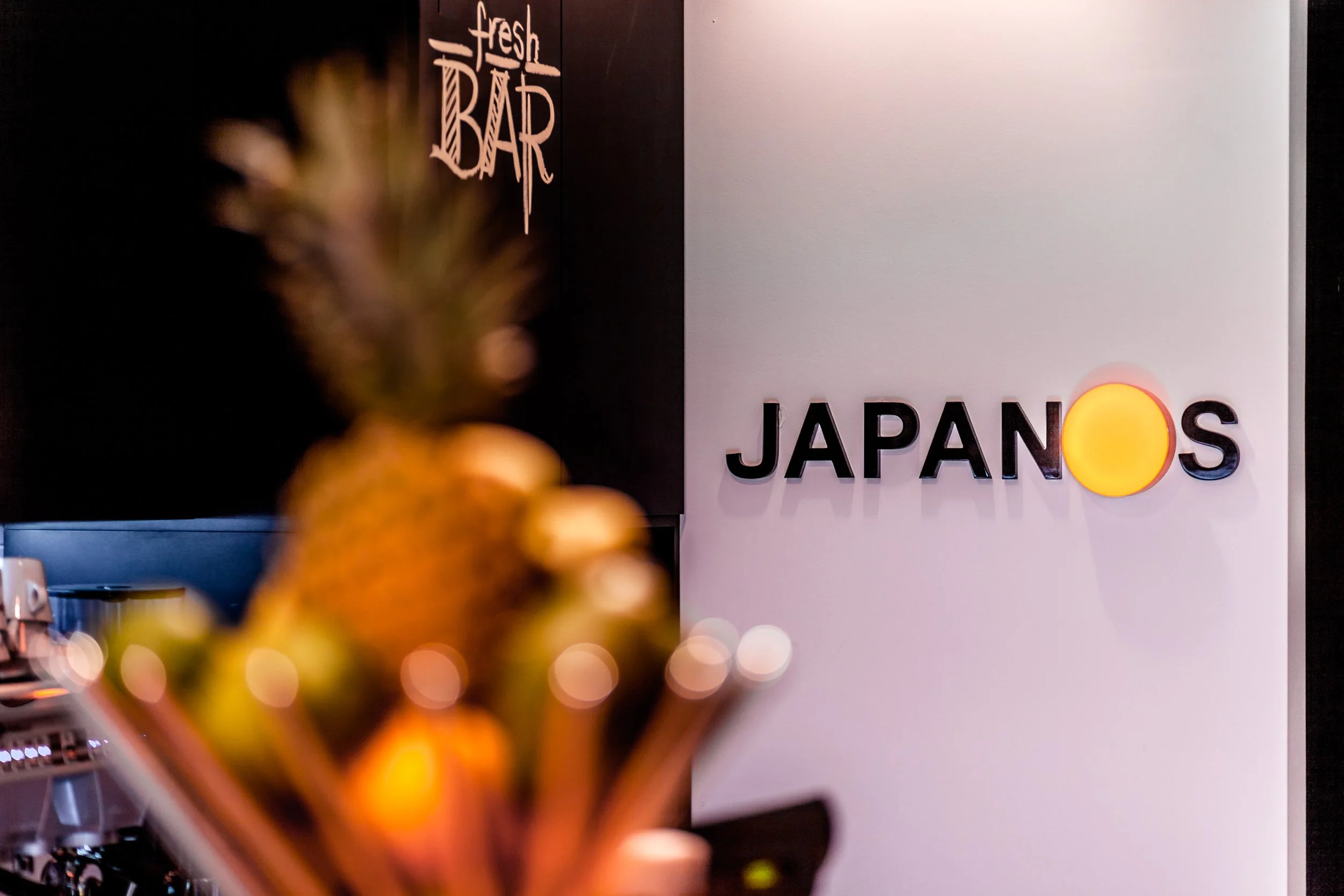 Blurred pineapple in foreground with a sign in the background that says 'fresh BAR' on a black wall and 'JAPANOS' on a white wall, with a yellow circle replacing the letter 'O'.