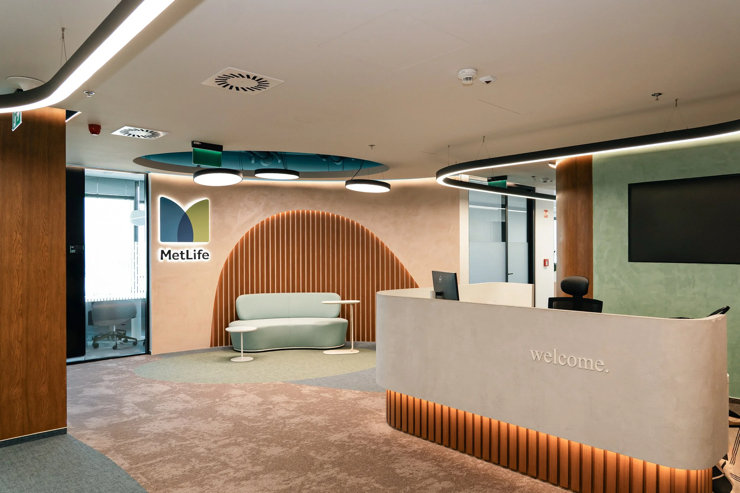Interior of a modern office reception area with a white curved reception desk, a black office chair, a green accent wall, a wall with a MetLife logo and a seating area with a light green sofa and small side tables, contemporary lighting, and various ceiling fixtures.