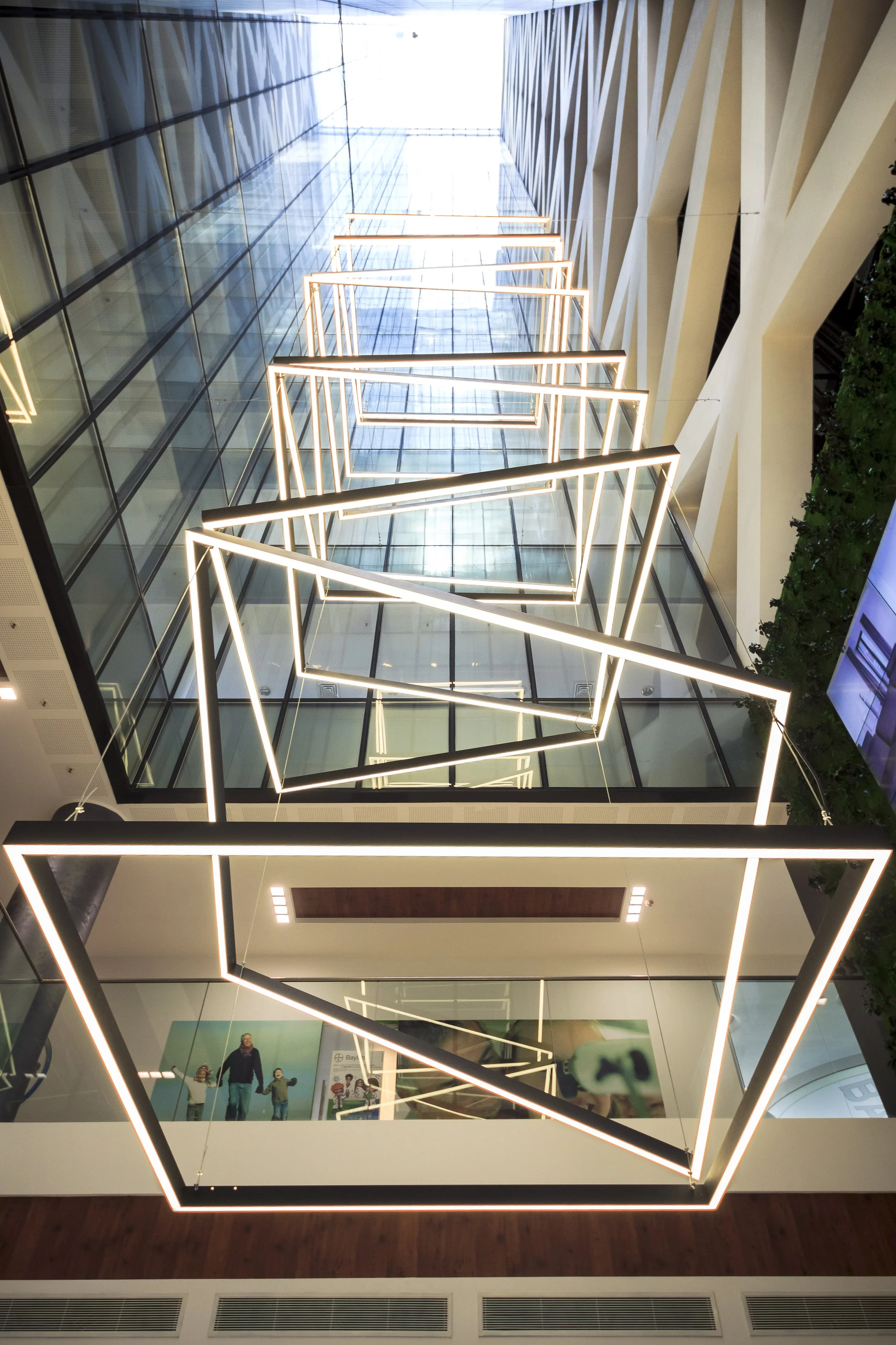 Modern building interior with geometric illuminated light fixtures hanging from the ceiling, capturing a view of the glass ceiling and the reflections below.