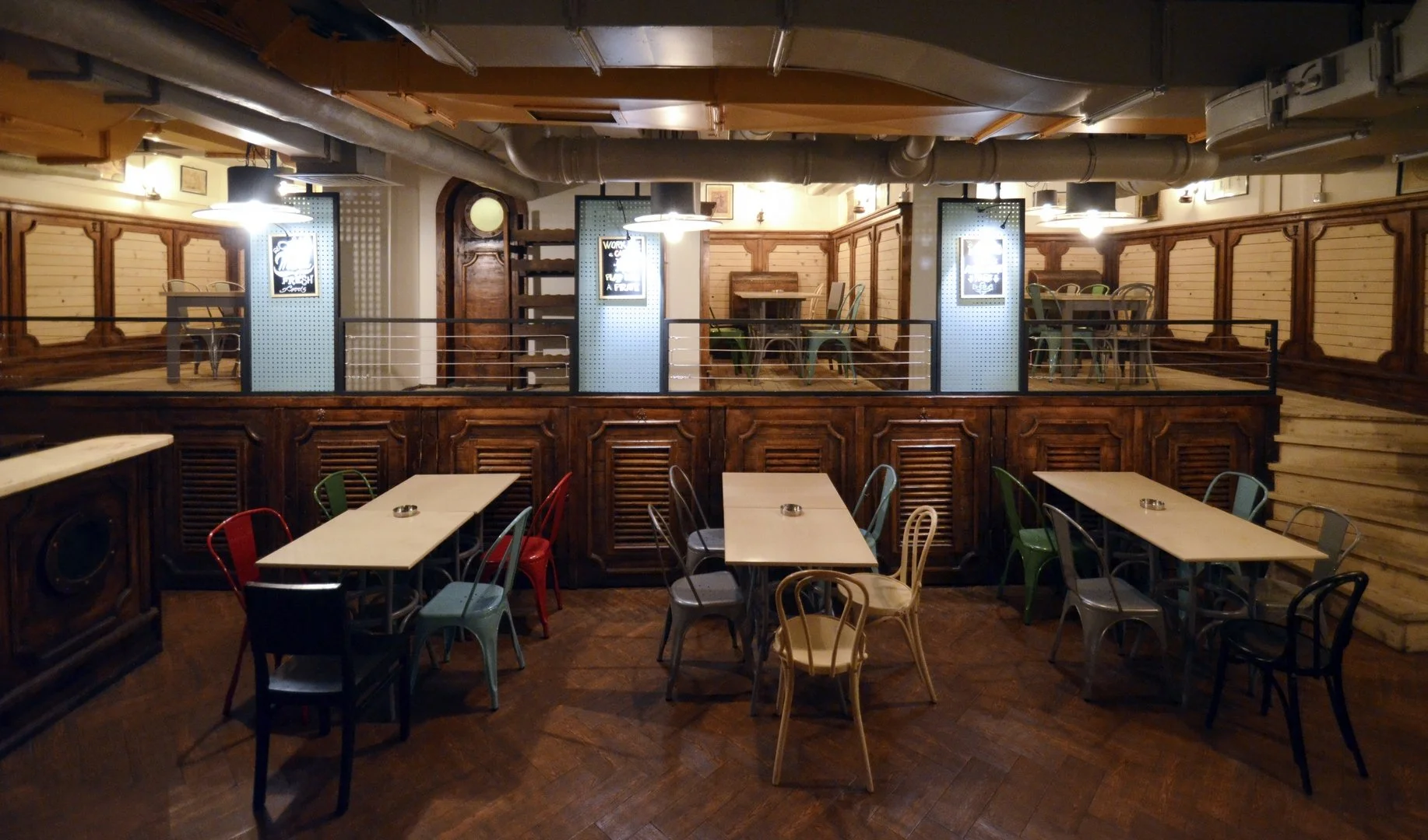 Cozy indoor restaurant with wooden decor, multiple tables and colorful chairs, upstairs dining area with similar setup, warm lighting, pipes and accents on ceiling.