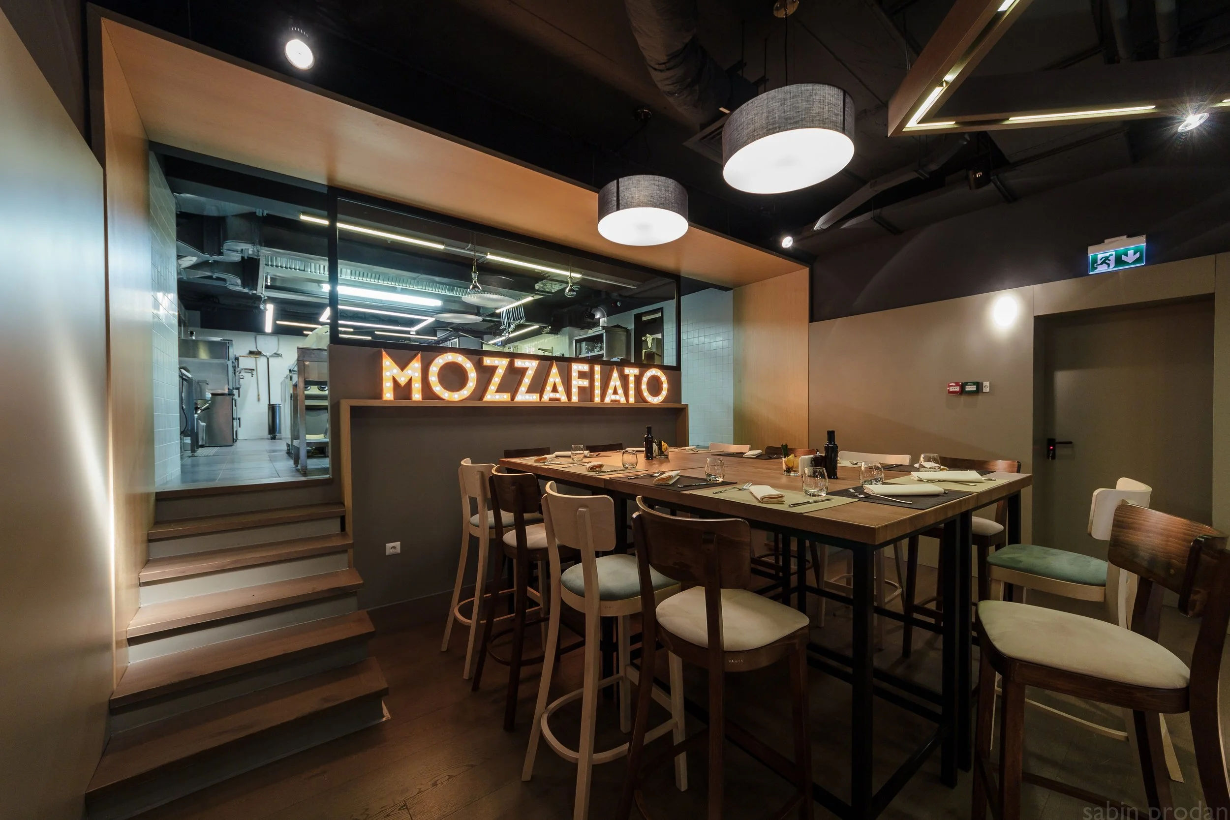Interior of a restaurant with a sign reading 'MOZZAFIATO' illuminated in lights, a long dining table set with glasses, plates, and utensils, and a view into the kitchen through a large window.