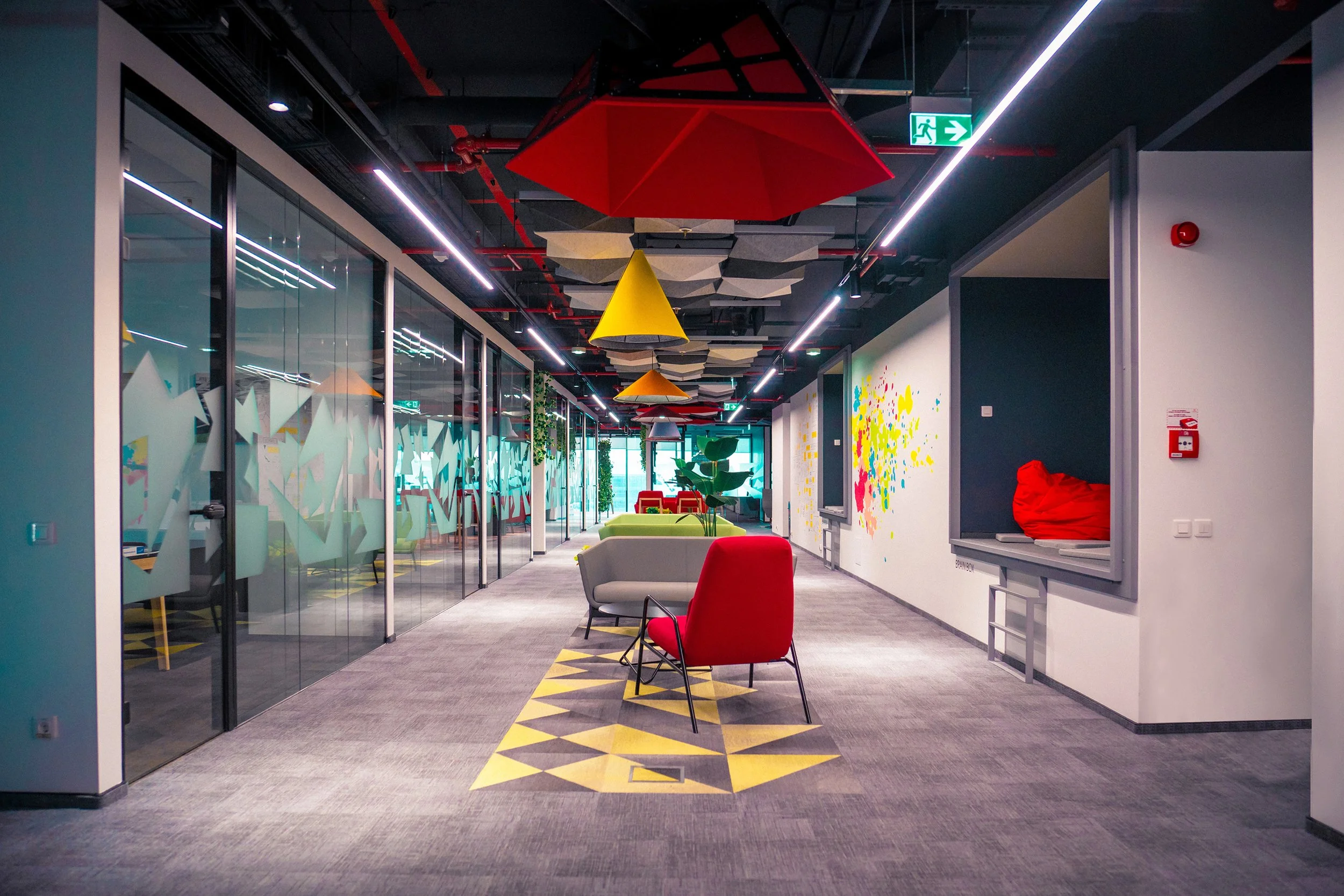 Modern office corridor with colorful chairs, large plants, and abstract wall art, illuminated by linear ceiling lights and decorated with hanging geometric fabric pieces and umbrellas.