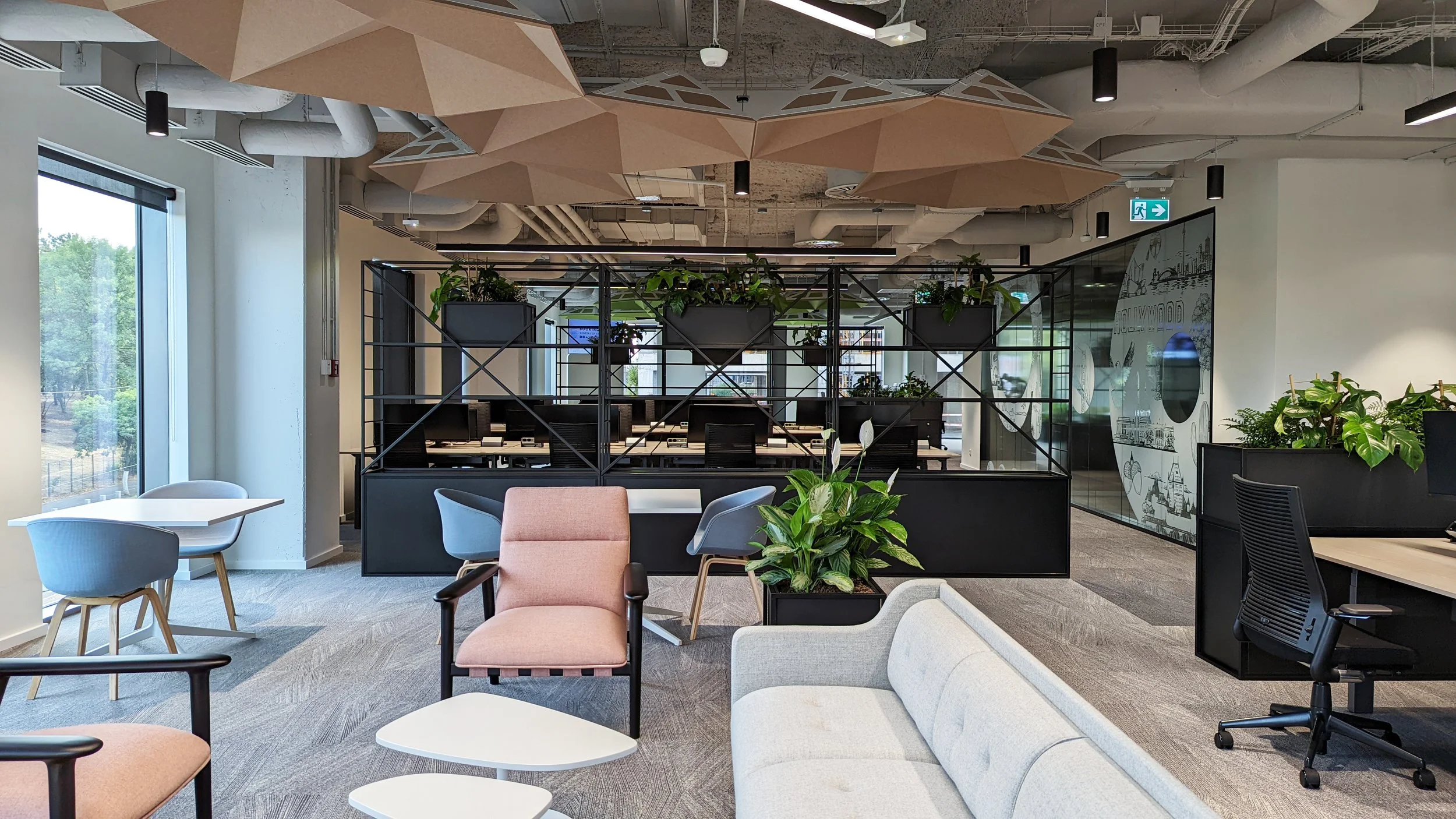 Modern office lounge with seating area, plants, and glass-walled meeting rooms, illuminated by large windows and ceiling lights.