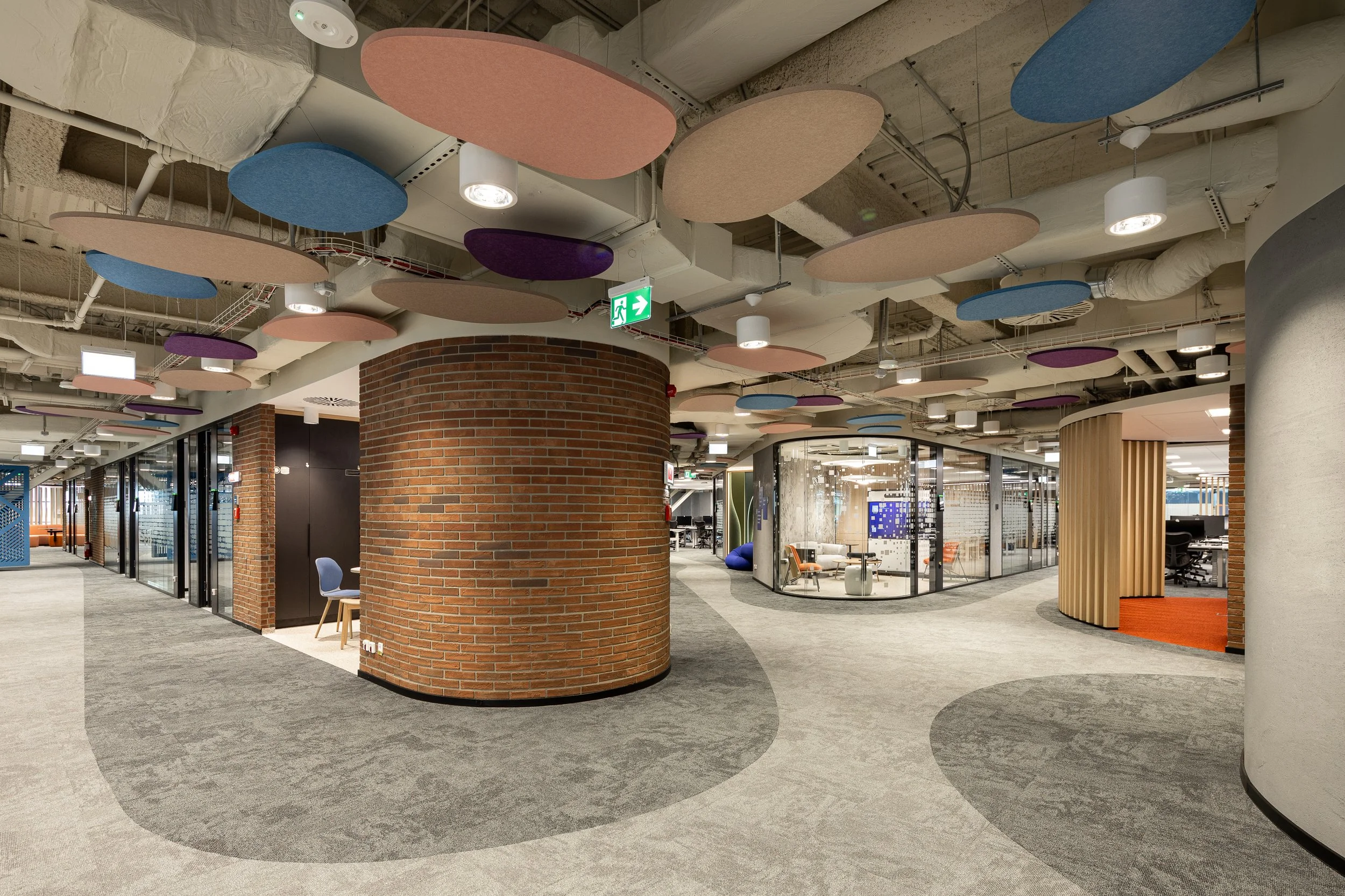 Modern office interior with brick walls, glass-walled meeting rooms, colorful ceiling panels, and a curved carpeted walkway.