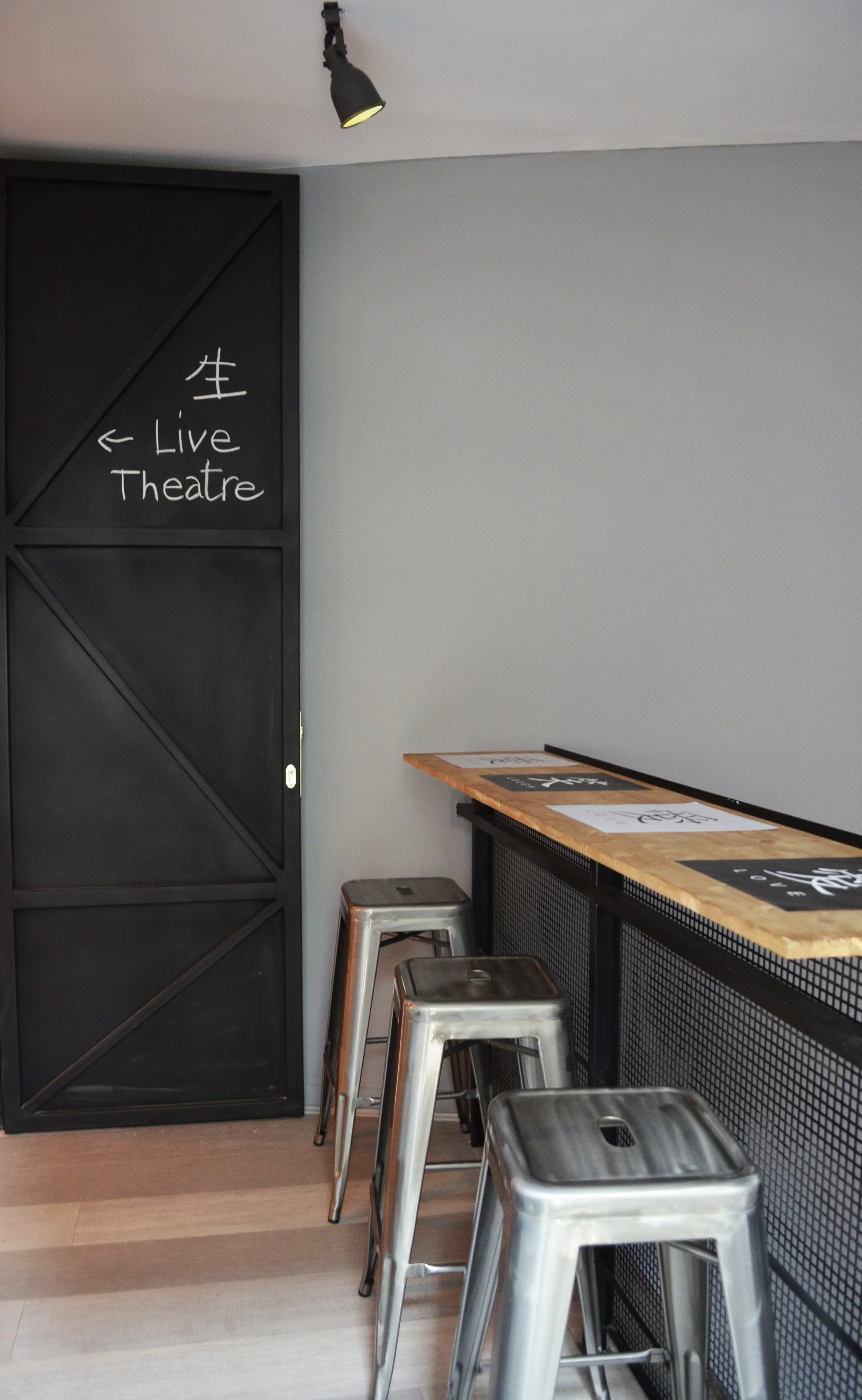 Interior of a modern cafe or bar with a high wooden counter, metal stools, and a blackboard sign indicating the direction to the live theatre.