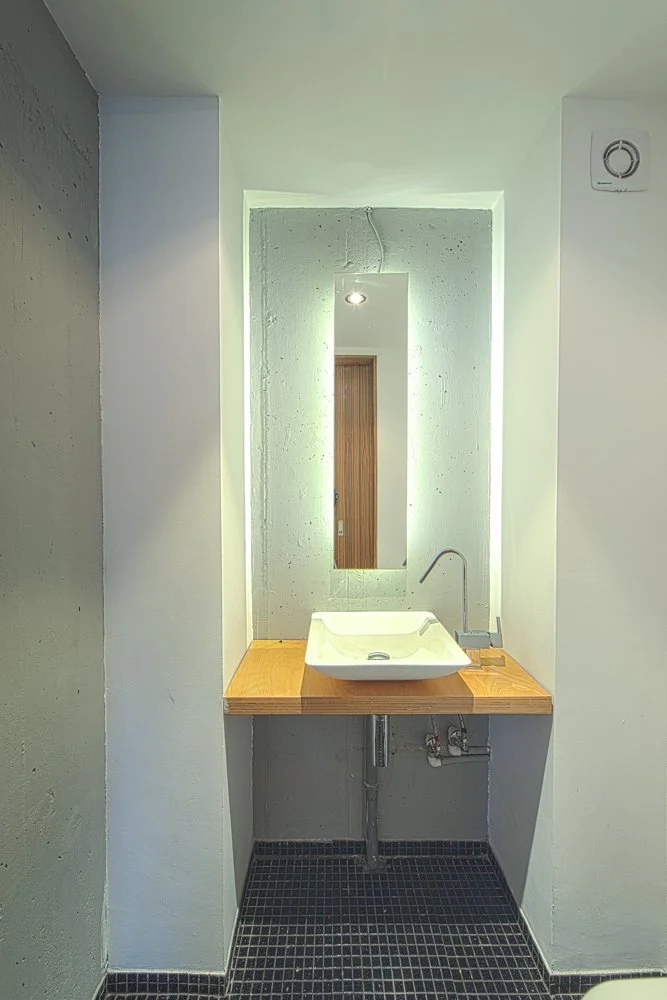 A minimalist bathroom with a rectangular mirror, a white vessel sink with a single-handle faucet on a wooden countertop, and black tiled flooring. The mirror is illuminated by a ceiling spot light, and a small wooden door is visible in the reflection.