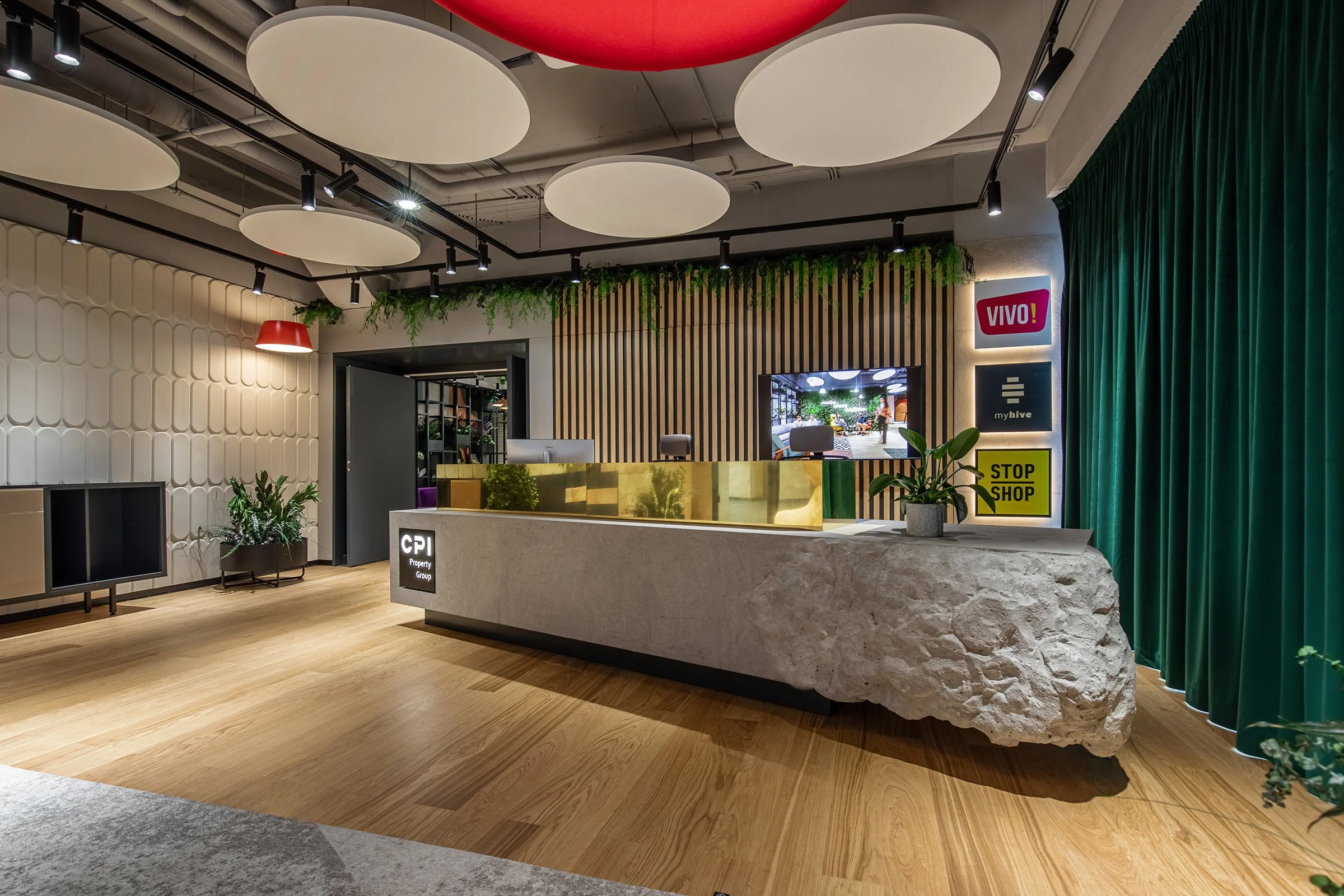 Modern hotel or office reception area with a stone front desk, green plants, wooden accents, and colorful signage. The ceiling has large circular light fixtures and acoustic panels.