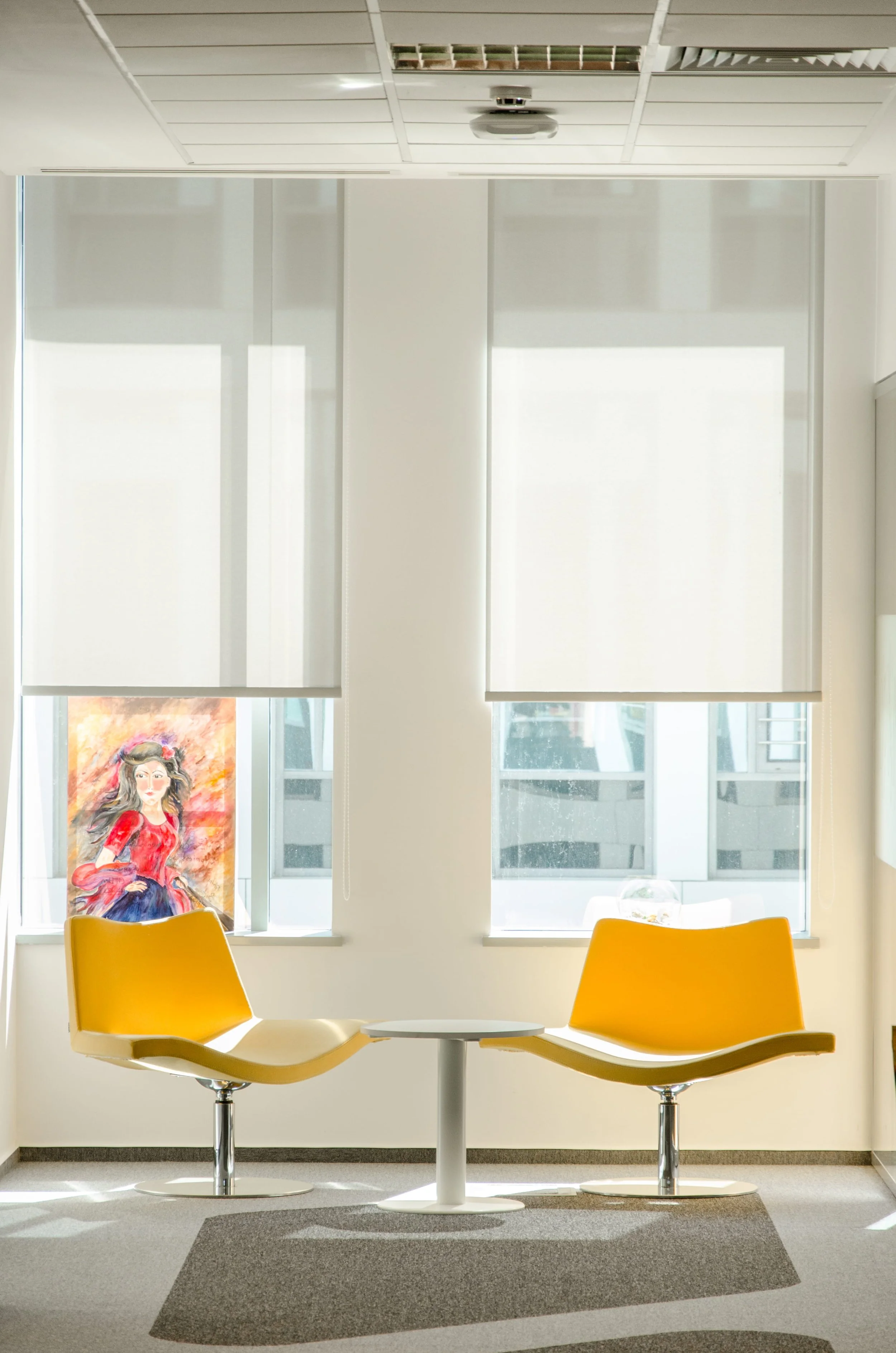 Empty lounge area with two yellow chairs facing each other and a small round table in between, positioned in front of two windows with white blinds and a poster of a woman in a red dress on the left window.