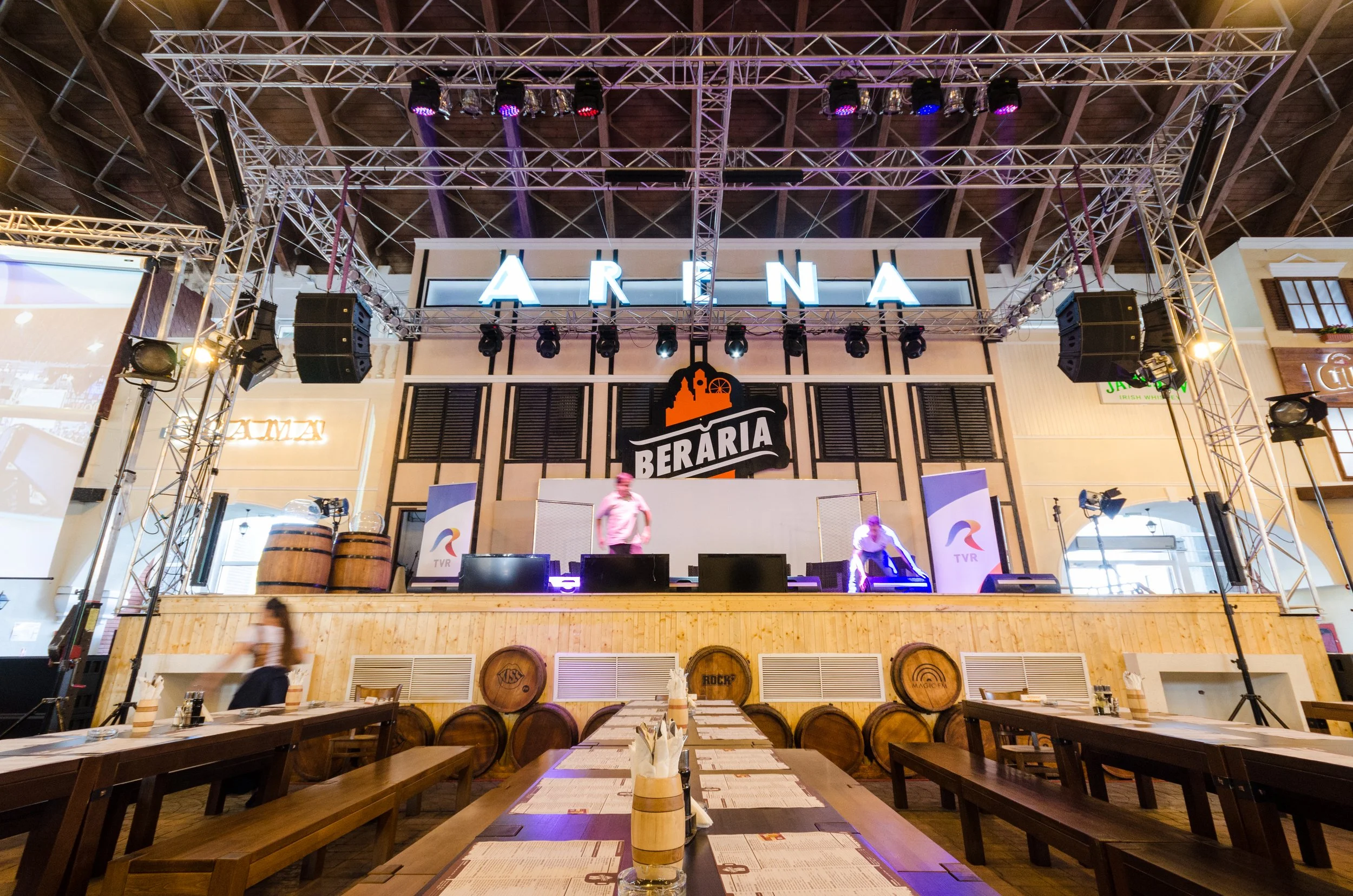 Interior of a restaurant or bar with a stage at the front, featuring a large sign that says 'ARENA' and a logo for 'BERARIA'. The stage is set with lights and speakers, and there are tables with menus and napkins in the foreground. Two people are working on or preparing the stage.