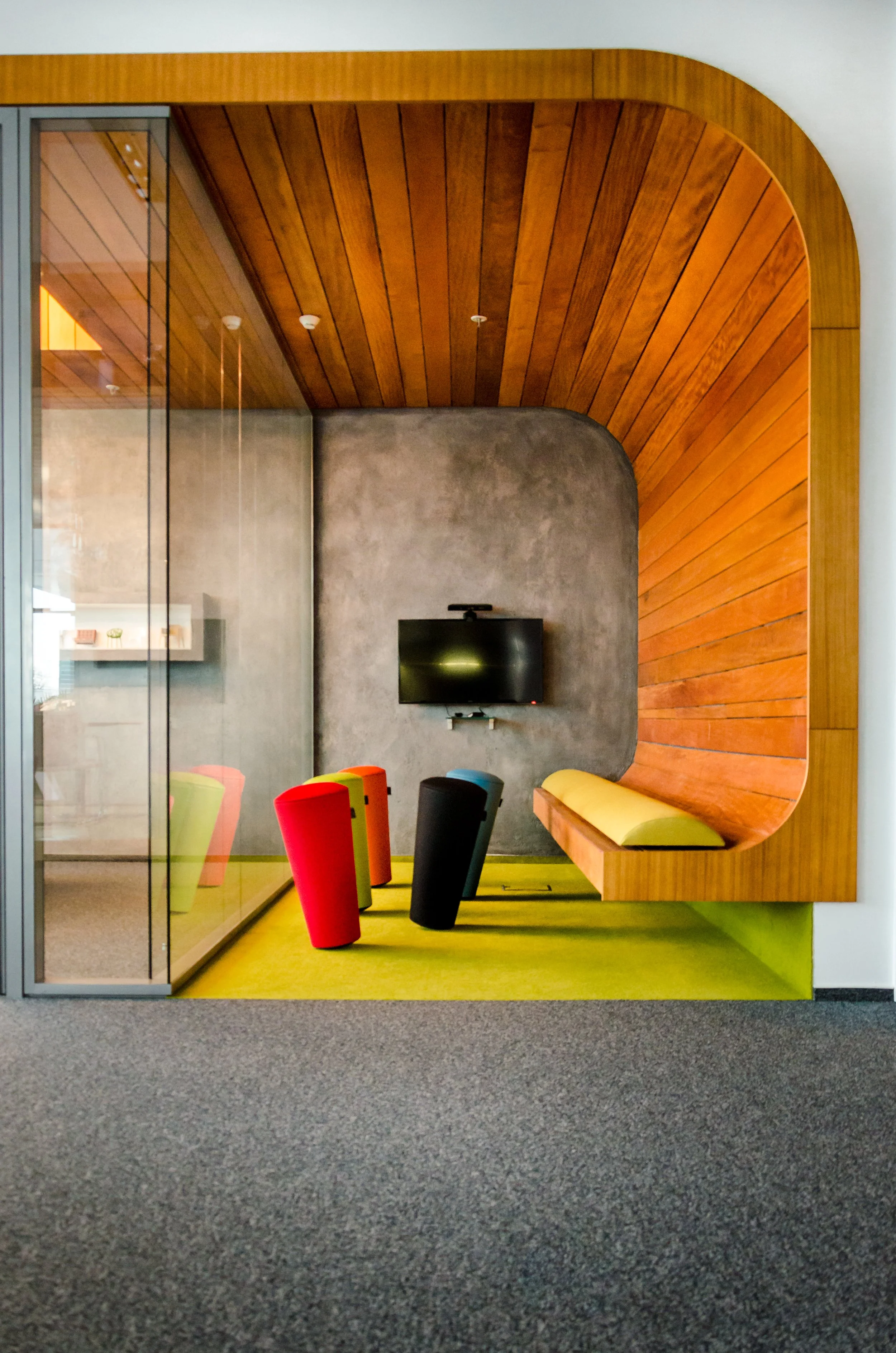 Modern office or lounge space with colorful chairs, wall-mounted TV, wooden and concrete walls, and a glass partition.