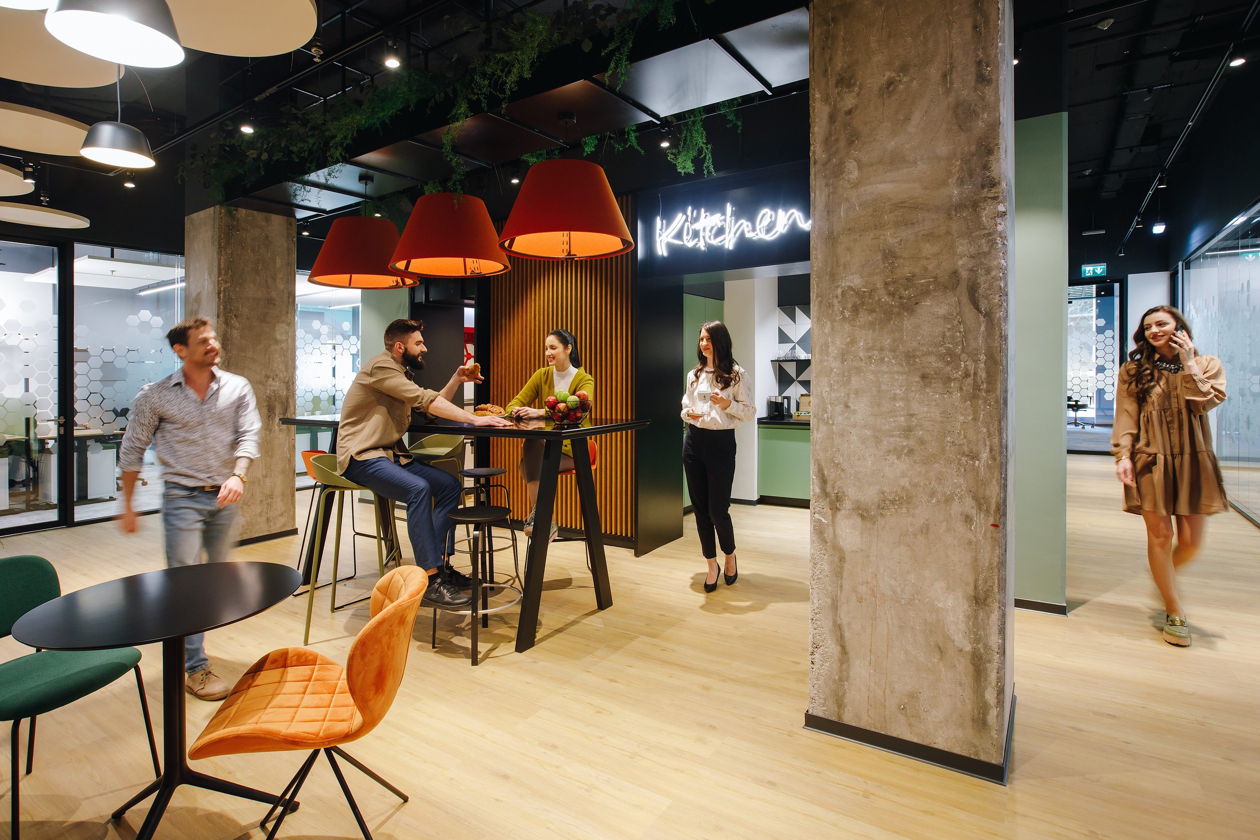 Office break room with people socializing and working, modern decor, illuminated 'Kitchen' sign, tables, chairs, and kitchen area in background.