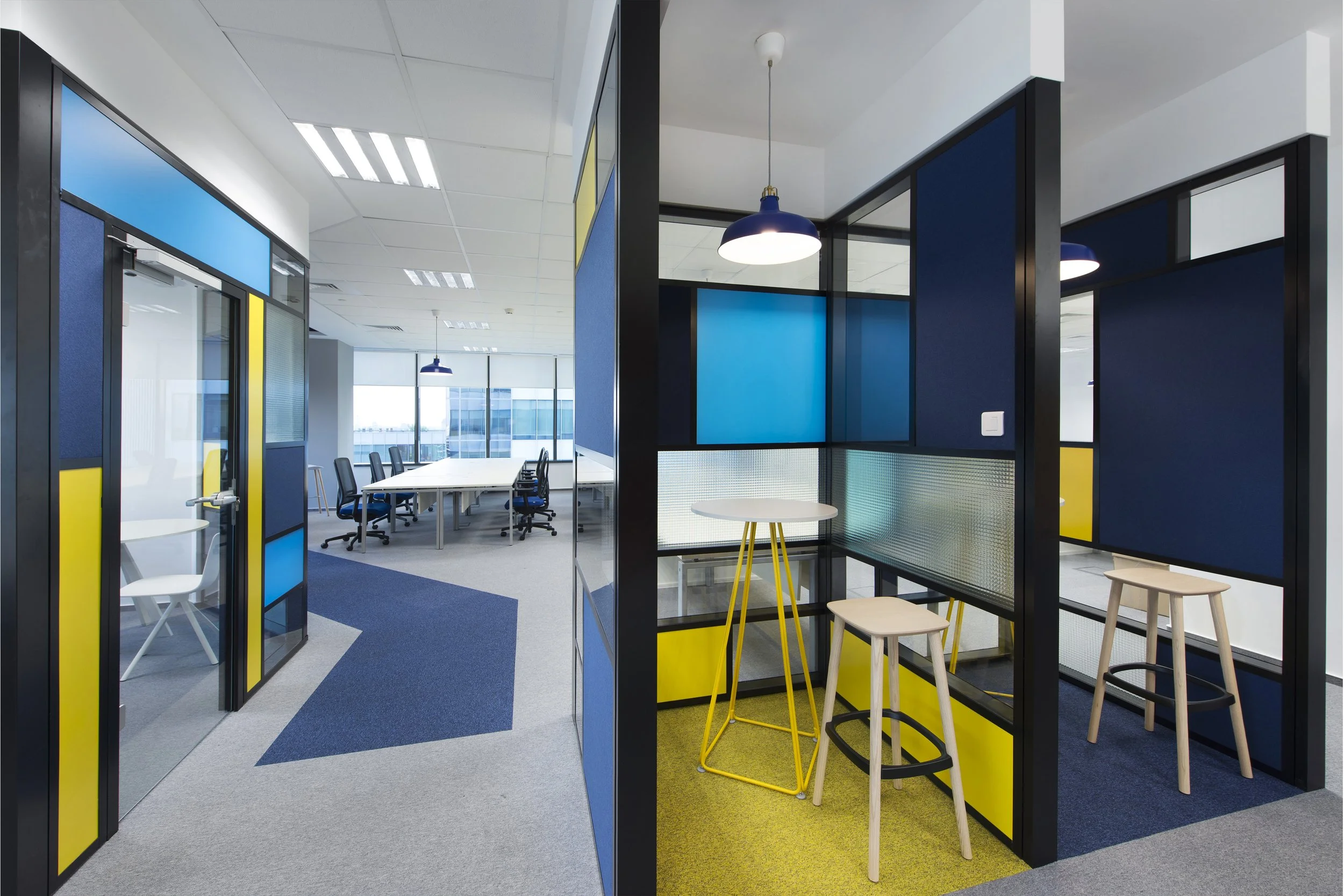 Modern office space with glass partitions, blue and yellow accents, white tables, black office chairs, and hanging pendant lights.