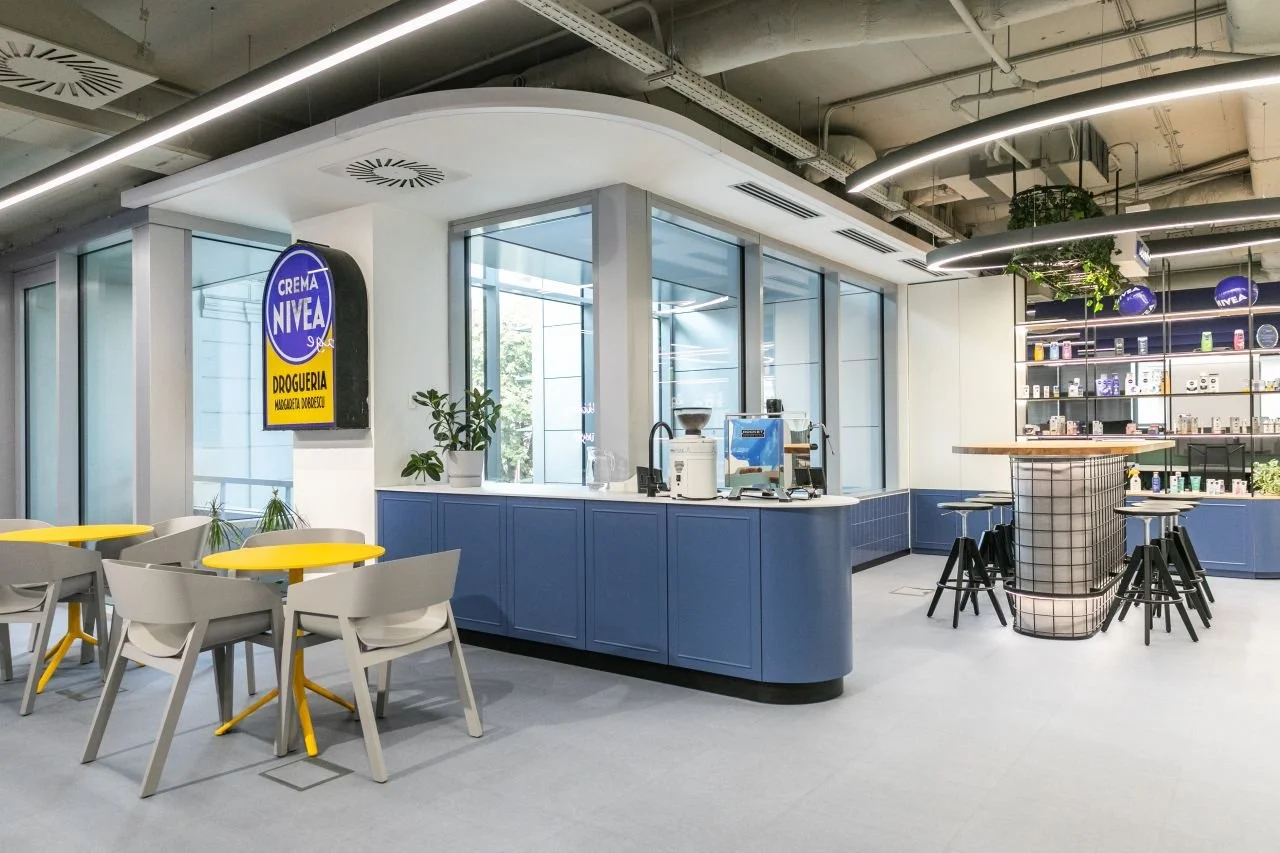 Modern coffee shop interior with blue and white color scheme, seating area with white and yellow tables, bar area with black stools, shelves with products, and large windows allowing natural light.