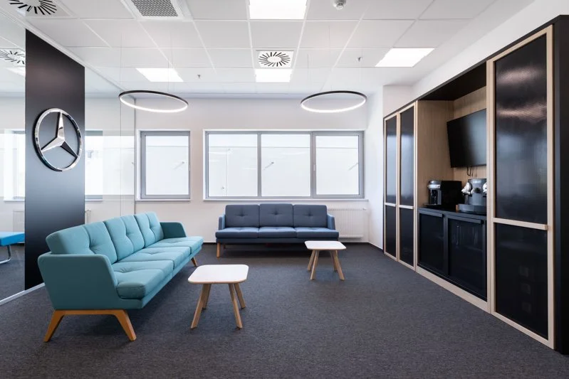 Modern office lounge with two light blue and dark gray sofas, three small wooden tables, large windows, a Mercedes-Benz logo on the wall, and a built-in coffee station with a TV.
