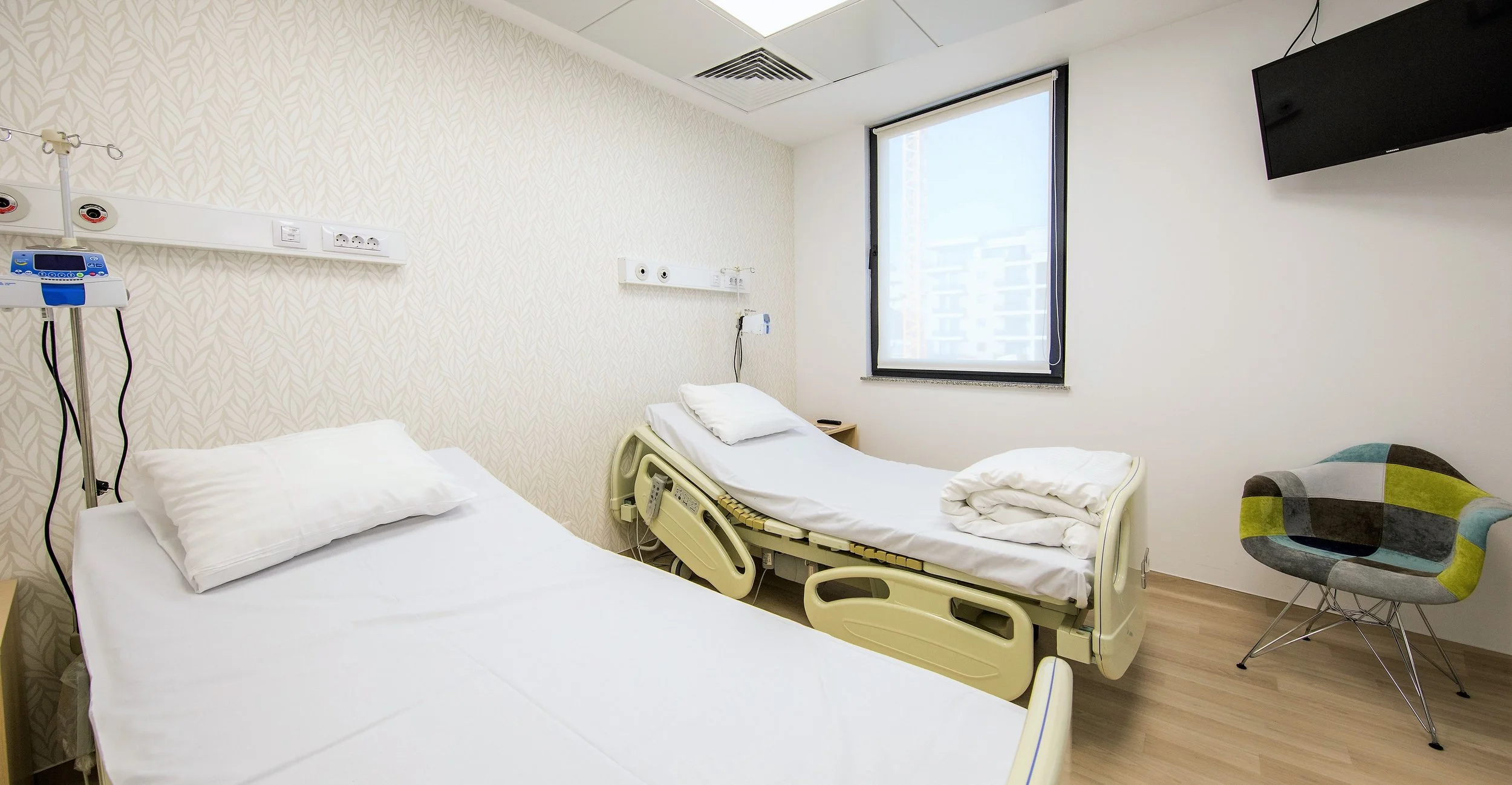 Hospital room with two beds, one adjustable, with white bedding and pillows, medical equipment on the wall, a window, a patterned armchair, and a wall-mounted TV.