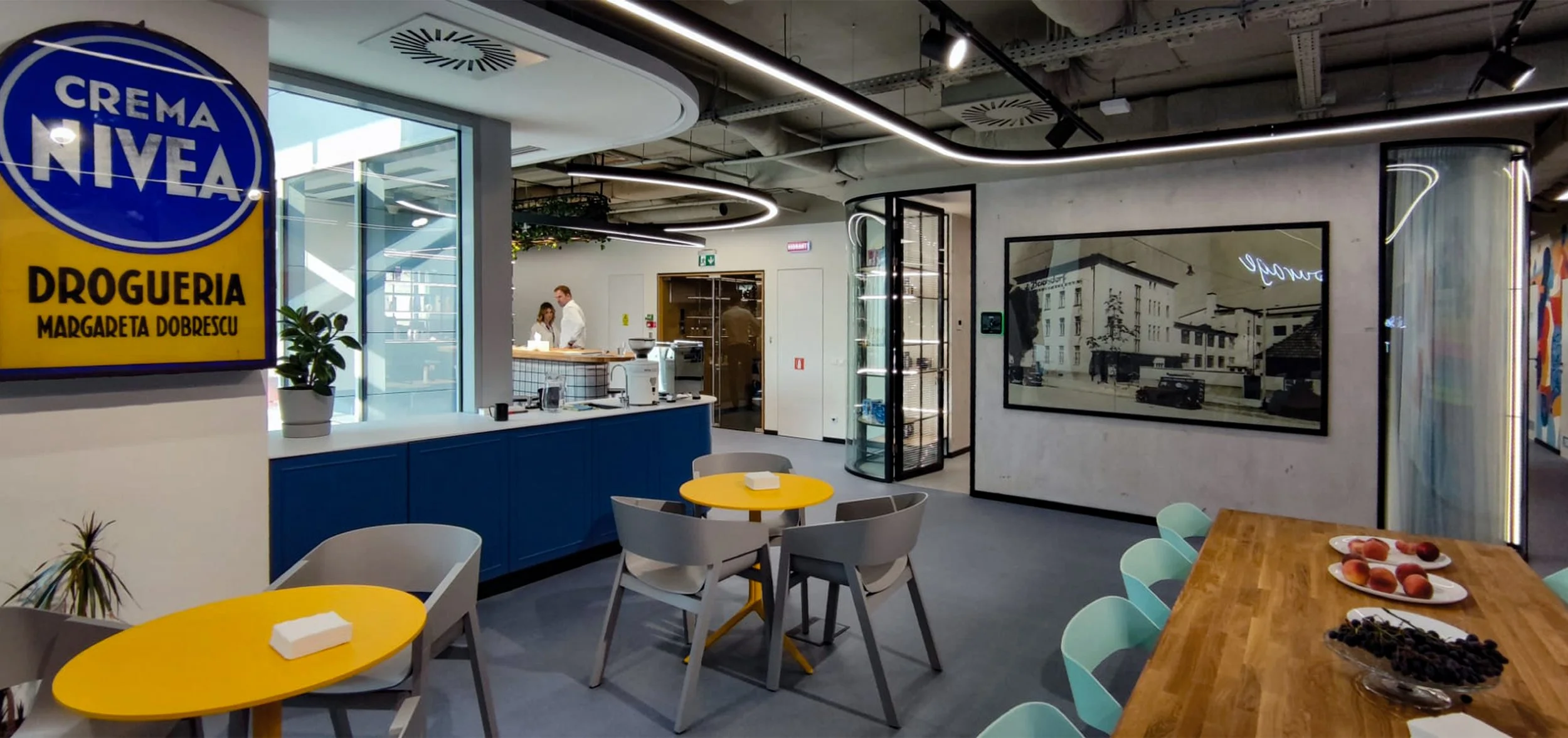 Modern office or café space with yellow and blue furniture, a coffee counter, and decorations. The area has a contemporary design with circular ceiling lights.