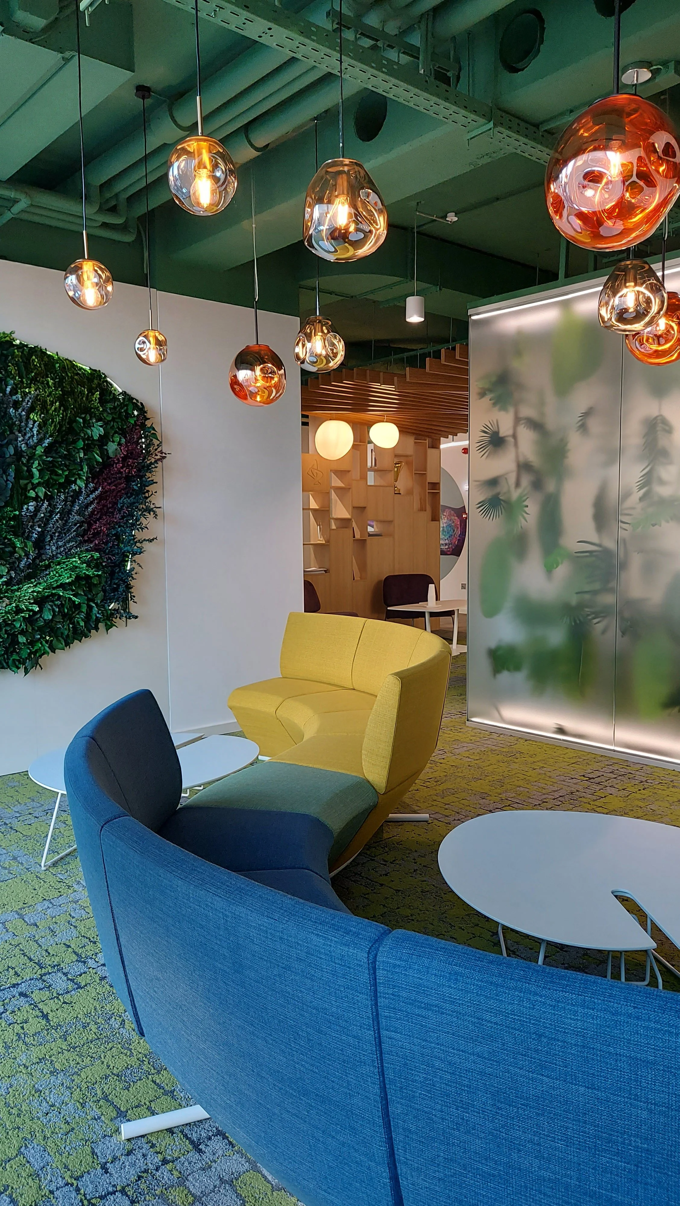 Modern lounge area with colorful curved chairs, small white tables, green patterned carpet, decorative wall art, and pendant lights hanging from a green ceiling.