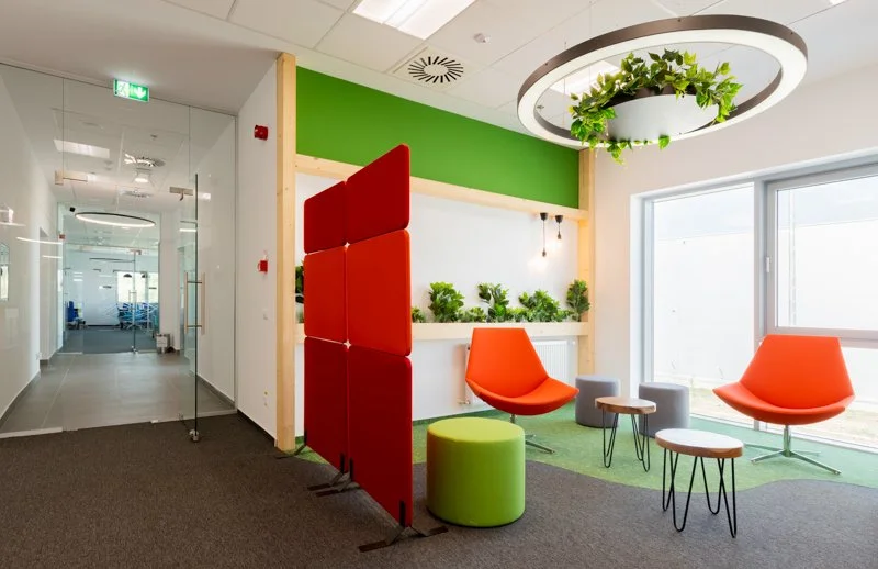 Modern office lounge area with orange chairs, green ottoman, small tables, and a hanging plant in a circular fixture.