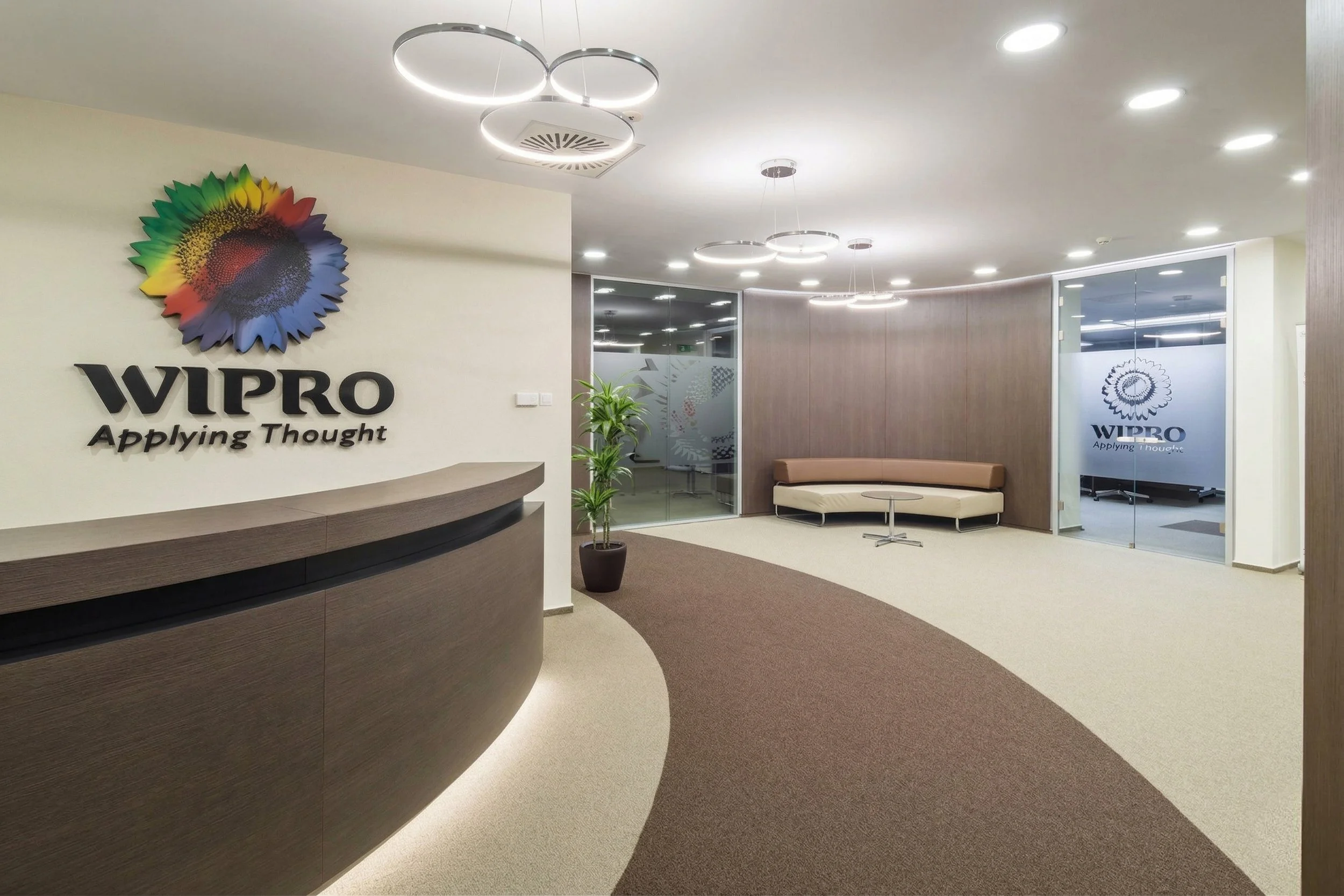 Modern office lobby with a reception desk, purple and brown carpet, seating area, and glass doors with the WIPRO logo.