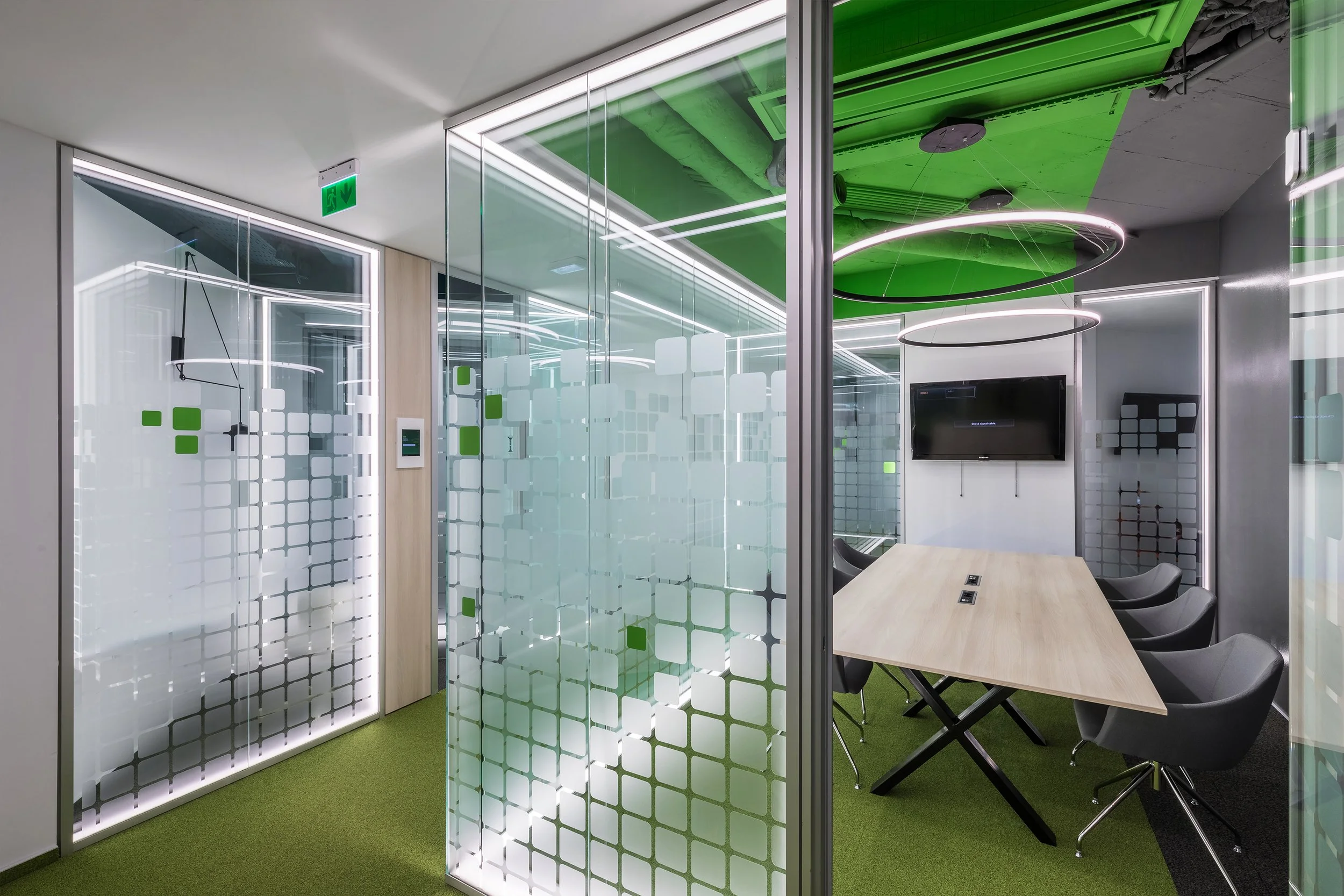Modern office conference room with glass walls, green ceiling accent, and a wooden table surrounded by black chairs. A large flat-screen TV is mounted on the wall, and there are circular LED light fixtures hanging from the ceiling.