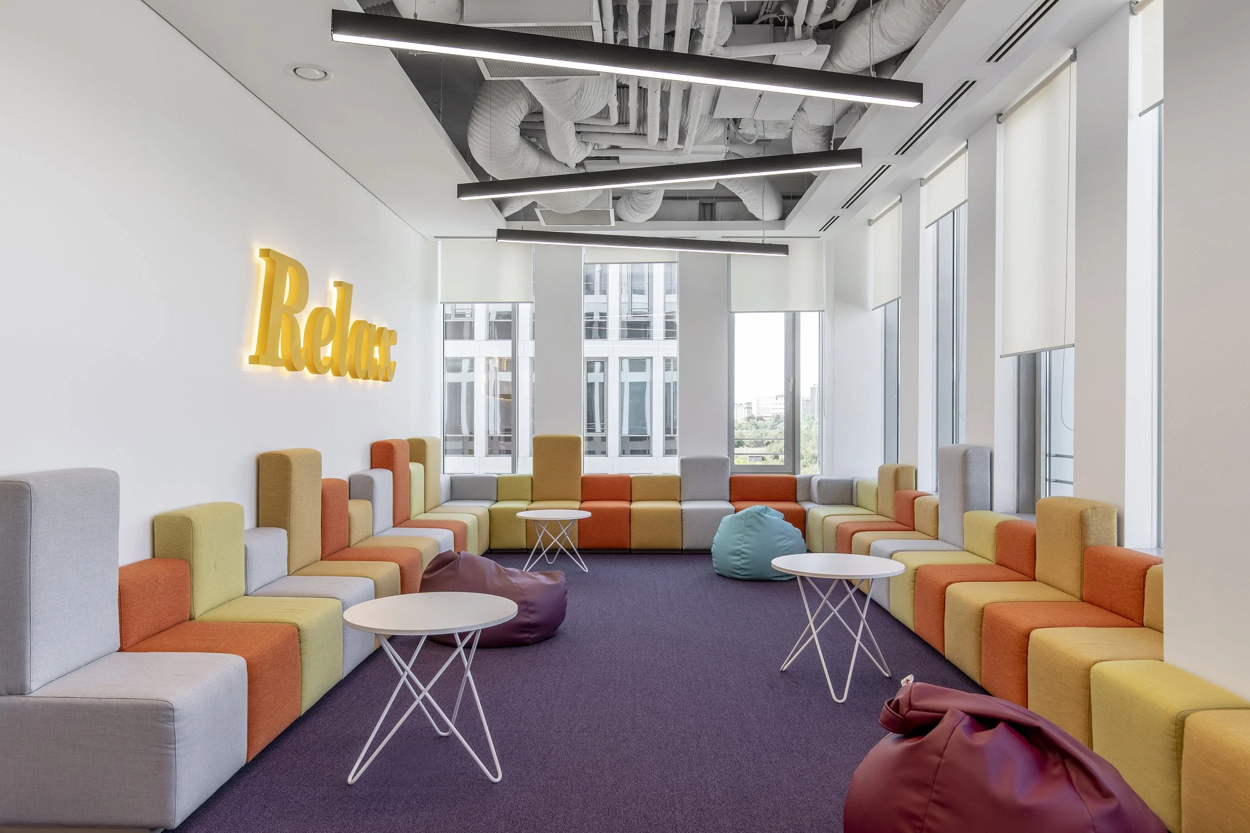 Modern bright lounge area with colorful modular seating, small round tables, bean bags, and a yellow neon 'Relax' sign on the white wall, with large windows and city view, ceiling with exposed ductwork and linear lighting.