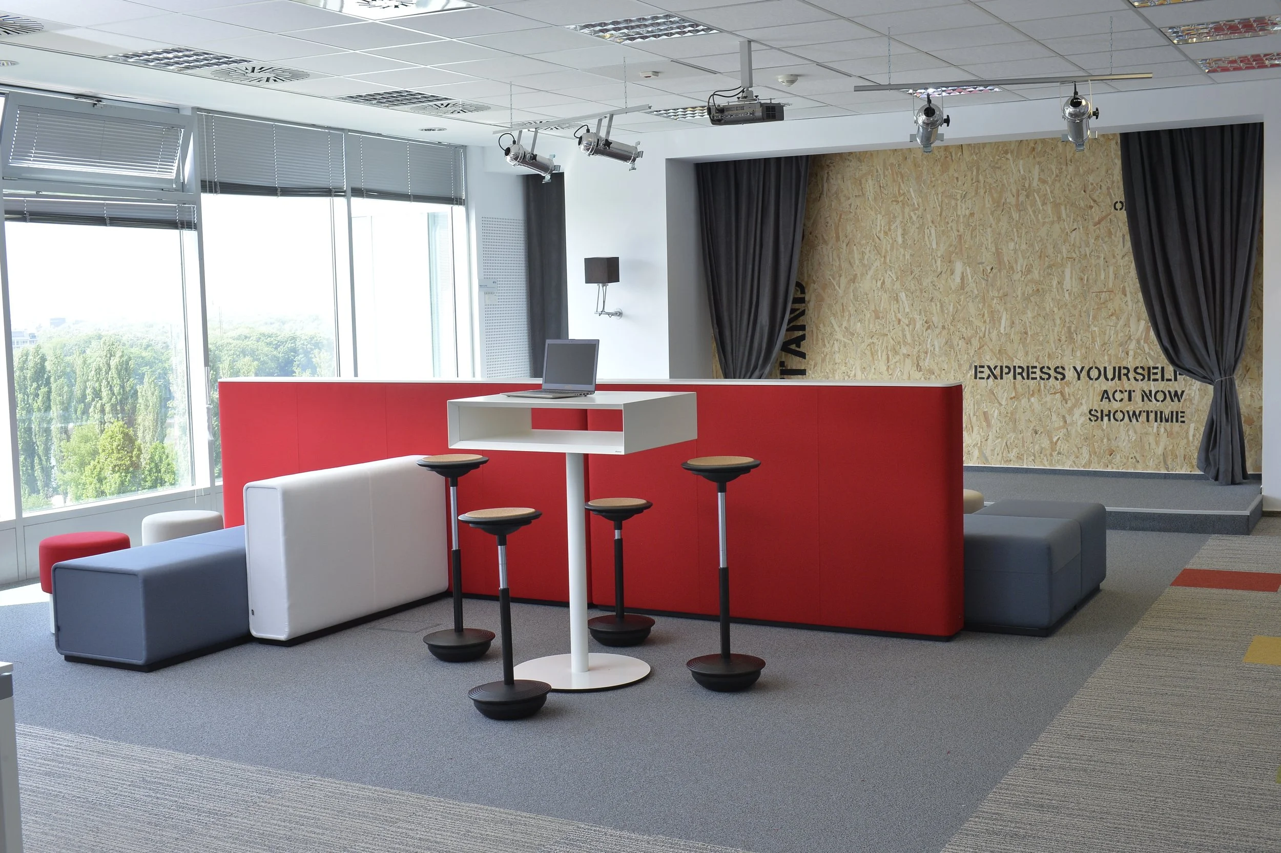 Modern office lounge area with large windows, a red partition, bar-height stools, a laptop on a white table, gray and white seating, and a stage with black curtains and a wooden backdrop with the words "Express yourself, act now, show time" written on it.