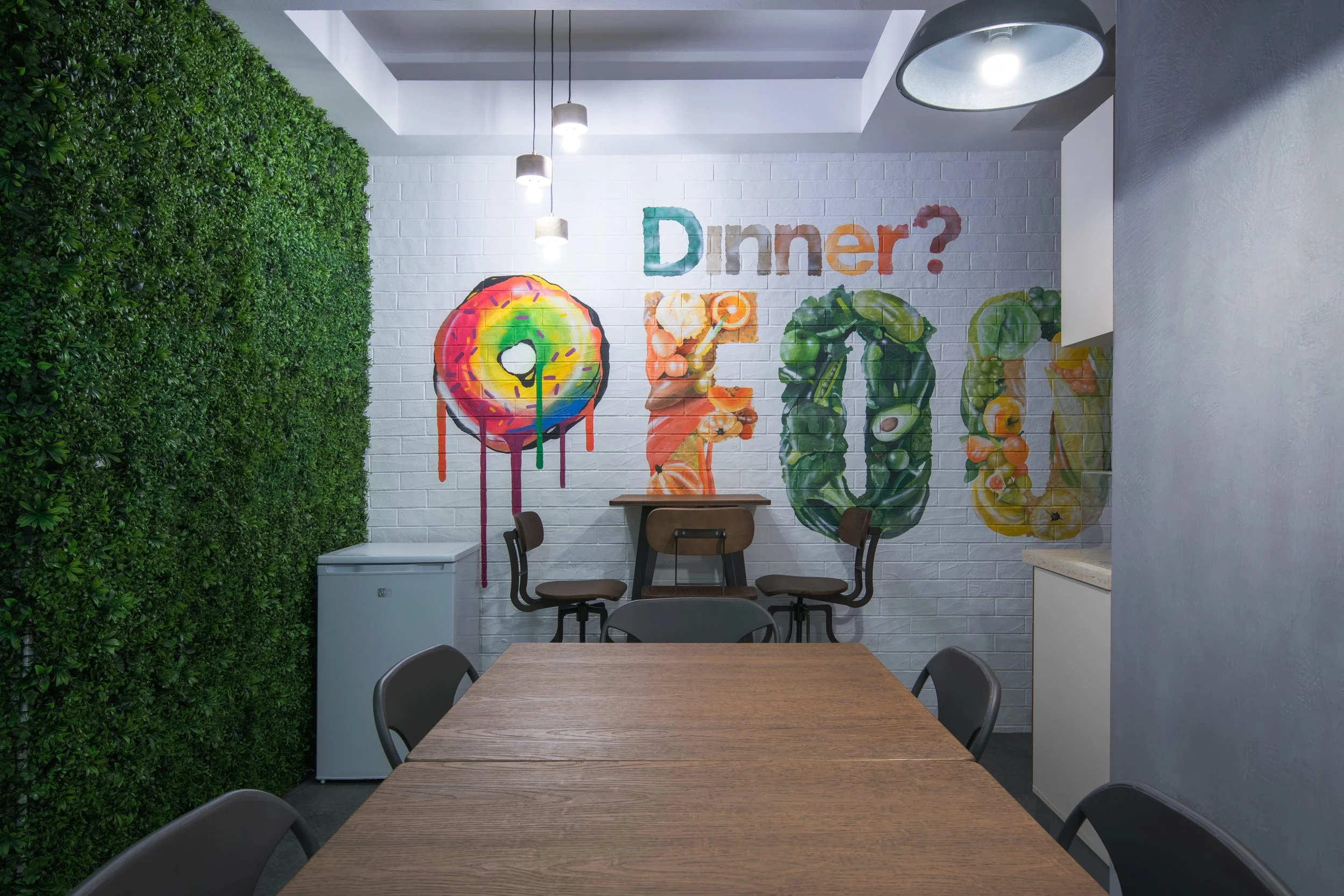 A dining area with a wooden table and black chairs. Blue and green artificial plants on a wall with a colorful mural. The mural spells 'Dinner?' with each letter painted as different vegetables and fruits, and a large colorful donut on the wall.