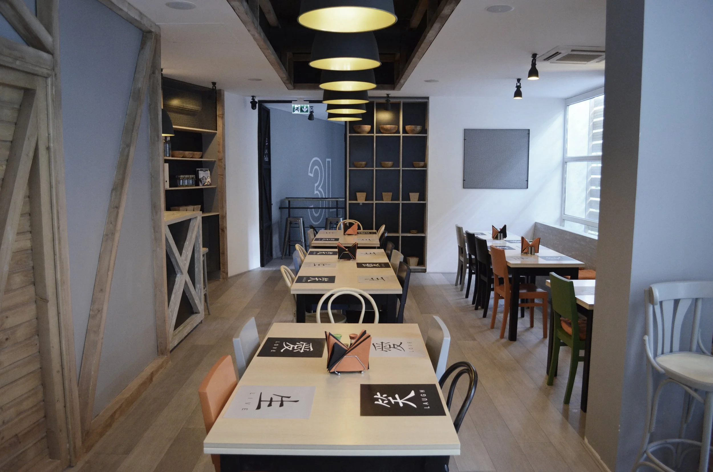 Interior of a modern restaurant with tables and chairs, some with napkin holders, a wooden wall, and a shelf with bowls and decorative items, illuminated by pendant lights and natural light from window