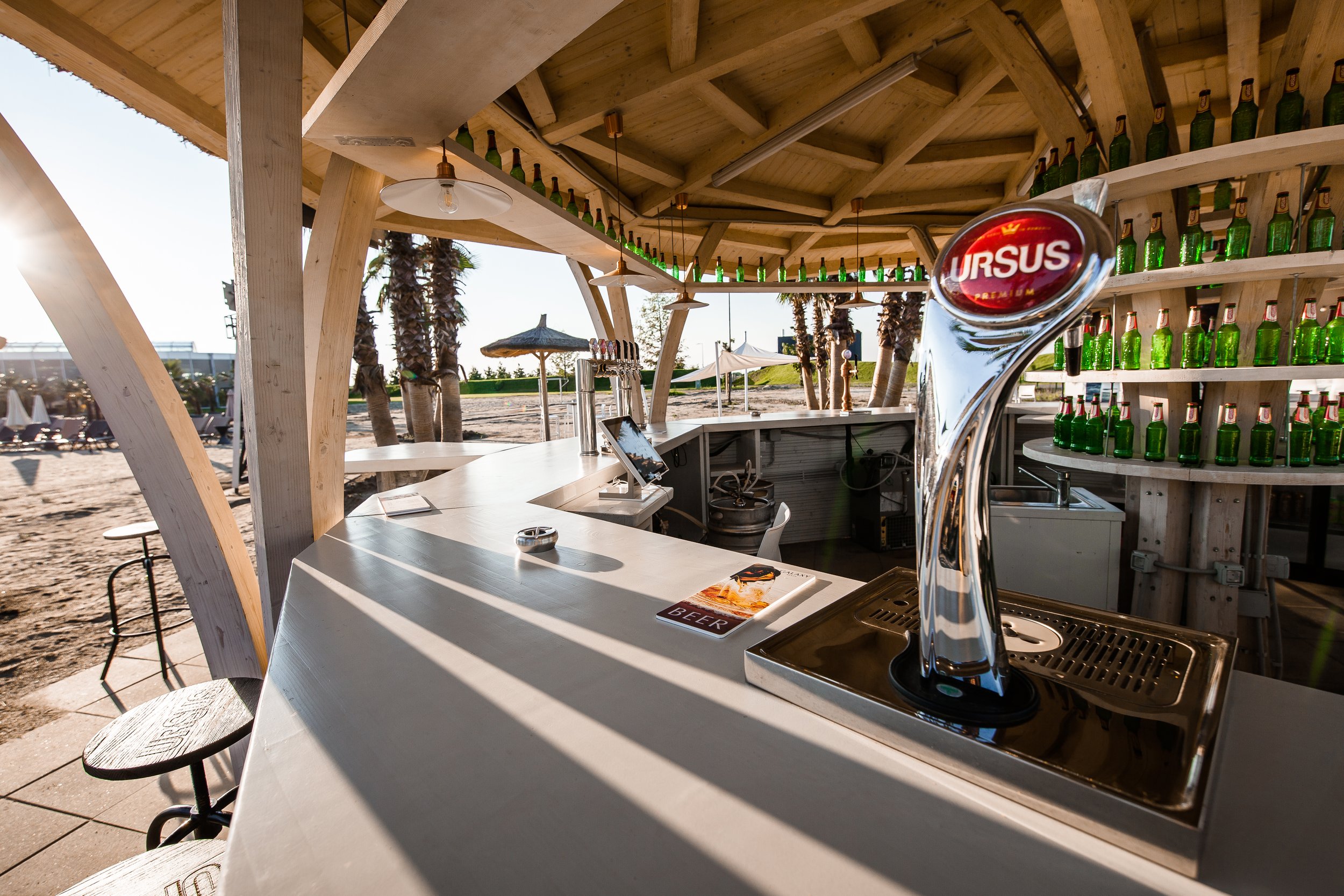 Beach bar with a white countertop, green bottled drinks on a shelf, a beer tap labeled "URSUS," and outdoor seating with palm trees and umbrellas in the background.