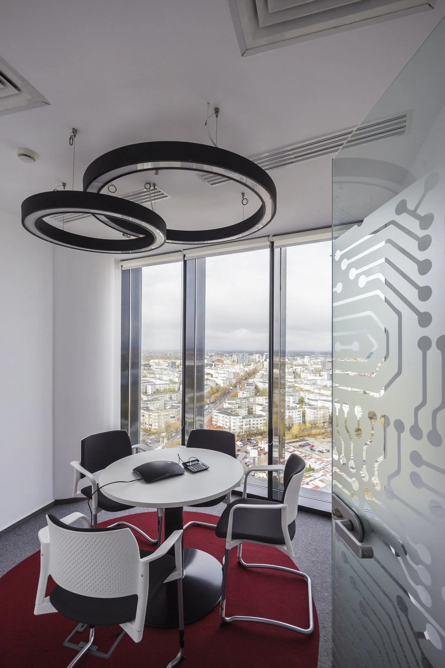 A modern conference room with a round table, four chairs, a conference phone, a calculator, and a large window with a city view. There's a decorative frosted glass wall with a circuit board pattern.