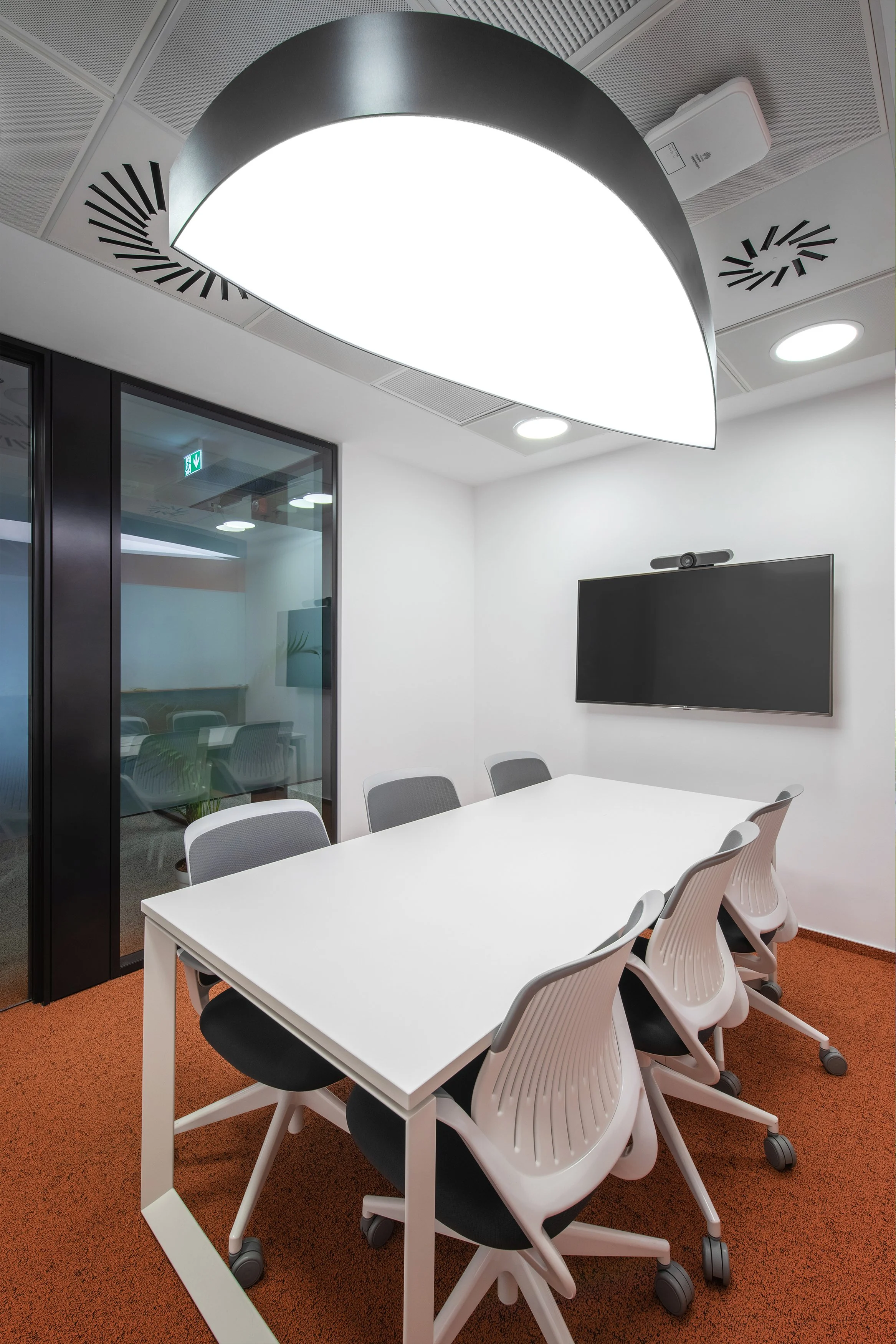 Modern conference room with a white table, six chairs, a large ceiling light, a wall-mounted screen, and a glass door leading to another room.