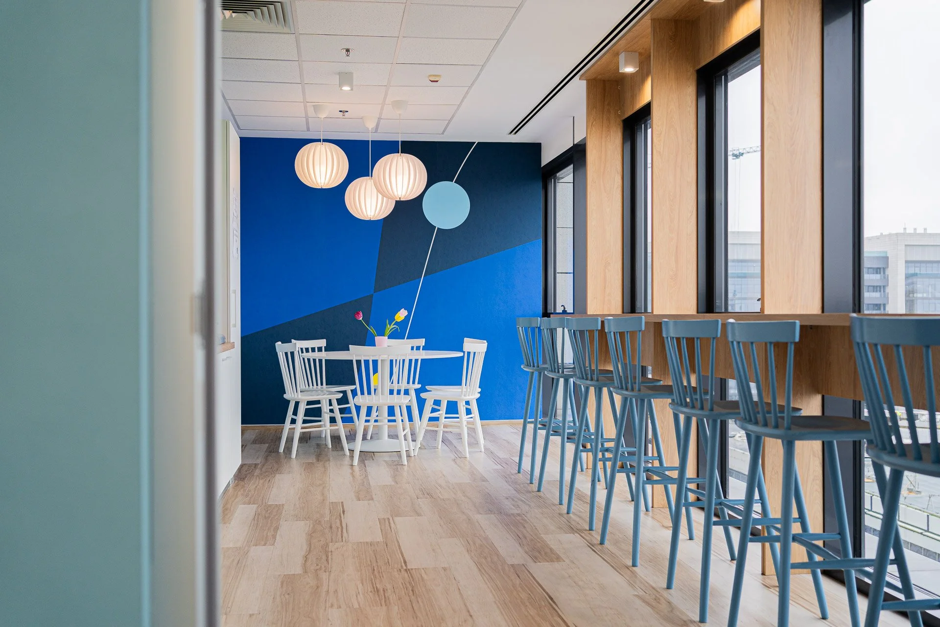 Modern cafe with high windows, blue accent wall, wooden paneling, bar seating, white round table with flowers and chairs, pendant lighting.