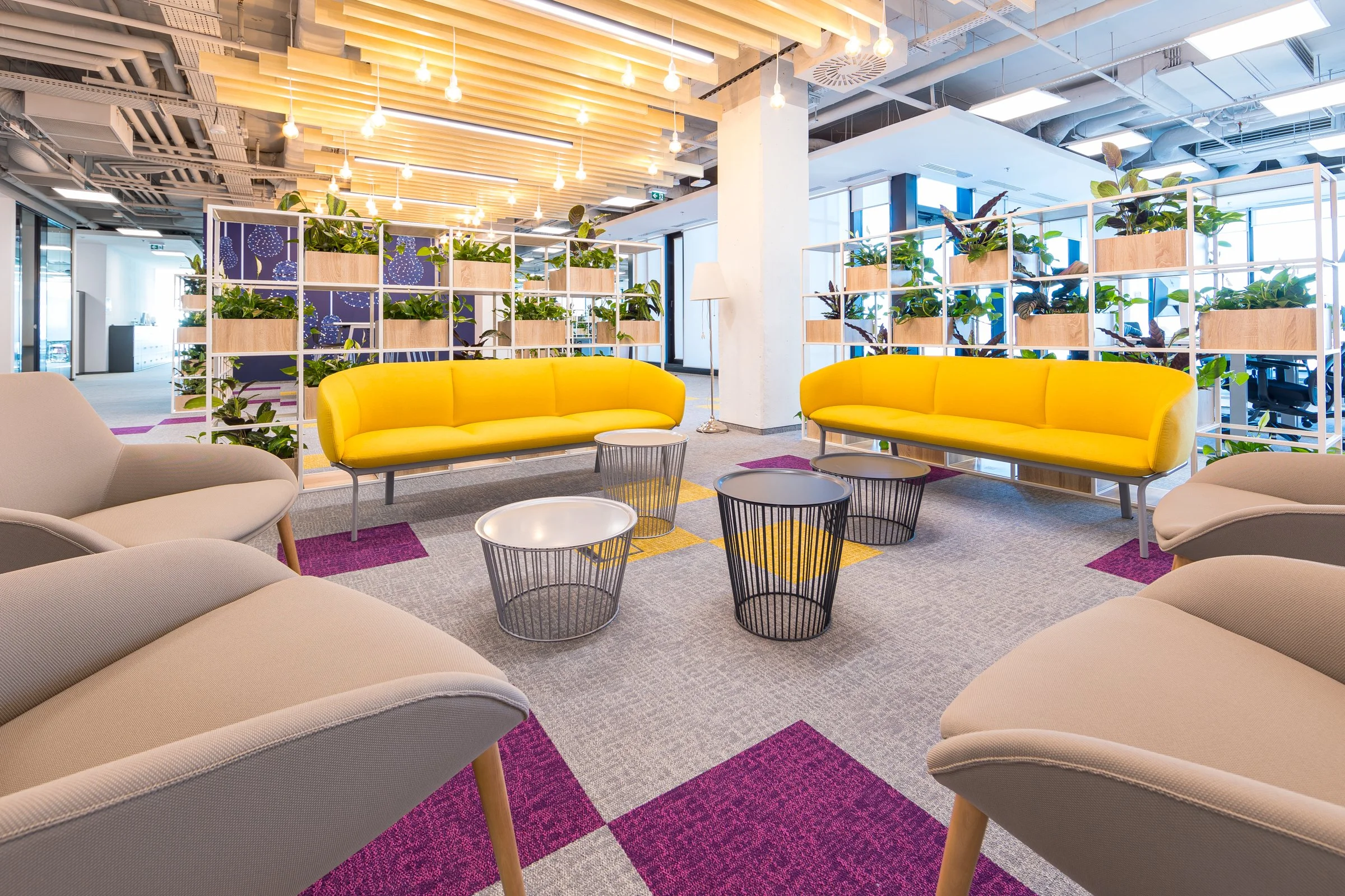 Modern office lounge area with beige and yellow seating, black wire coffee tables, and decorative white shelves with plants. Bright lighting and colorful carpet tiles create a welcoming atmosphere.