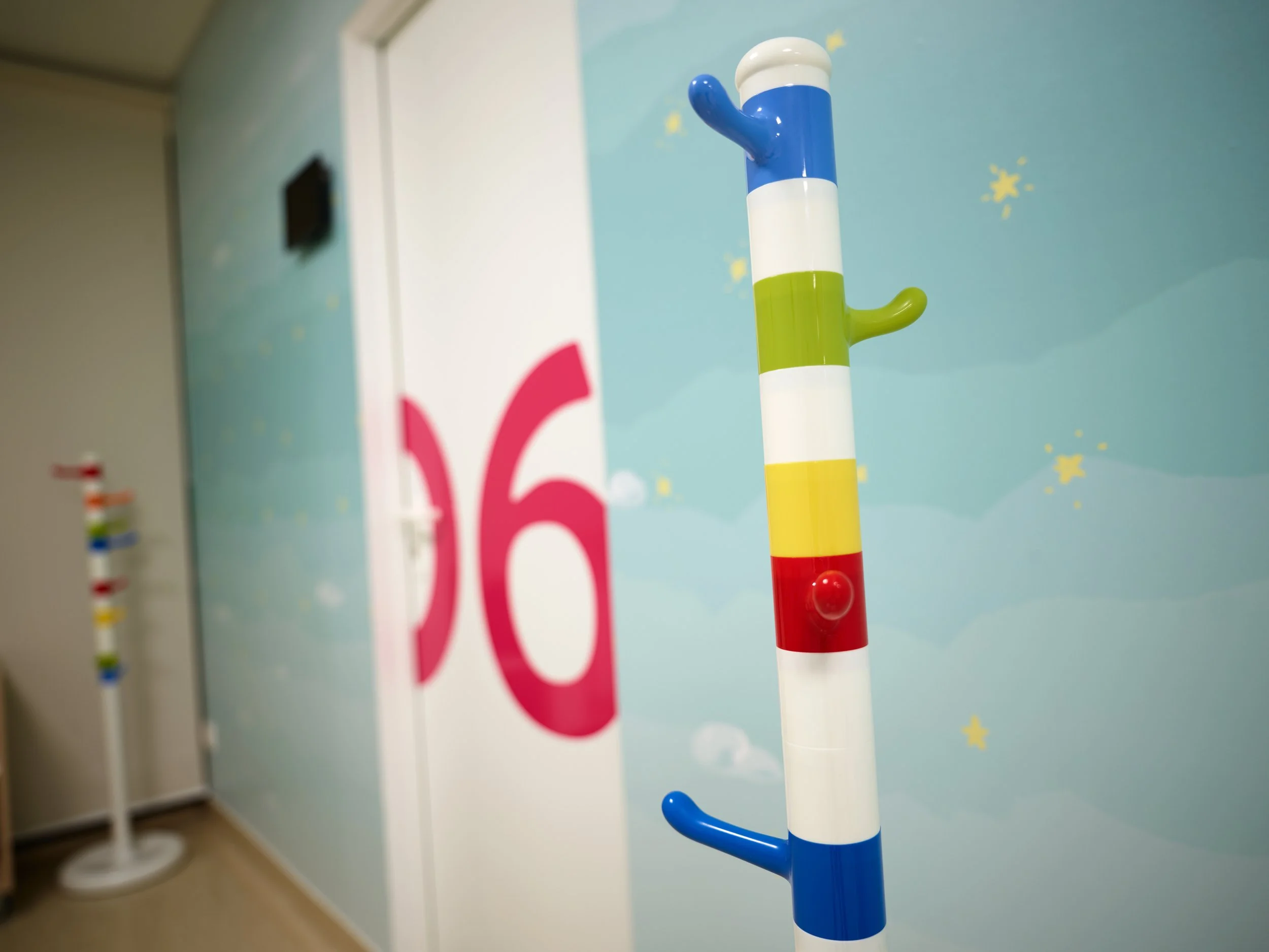 Colorful coat rack with sections in blue, white, yellow, and red, mounted on a light blue wall with yellow stars and clouds, and a large pink number 6 in the background.